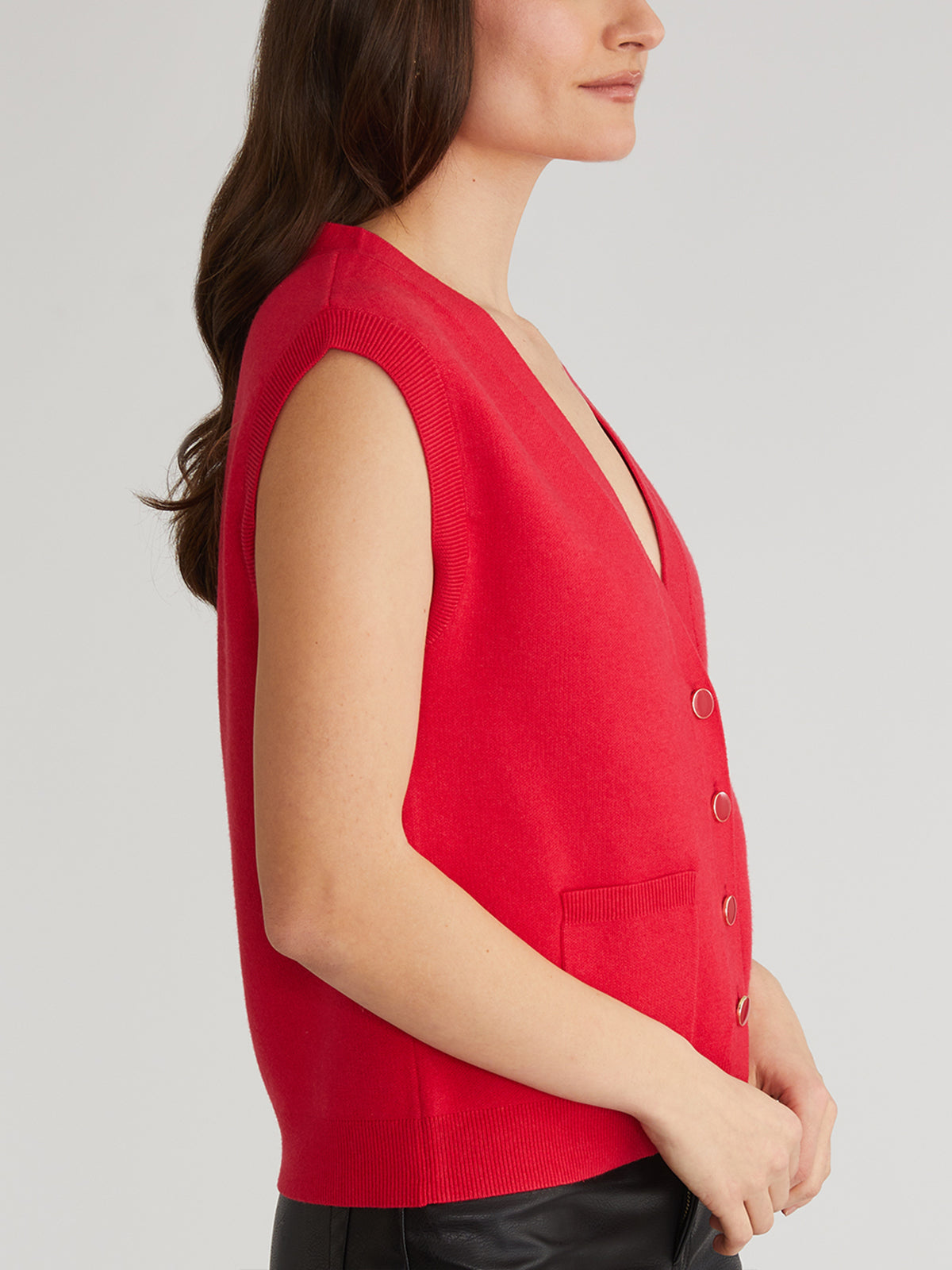 Wearing a chic 525 America red sleeveless button-down cardigan with a V-neck, two pockets, perfect for layering. The vest enhances their long dark hair as they pose from the side against a plain white background.