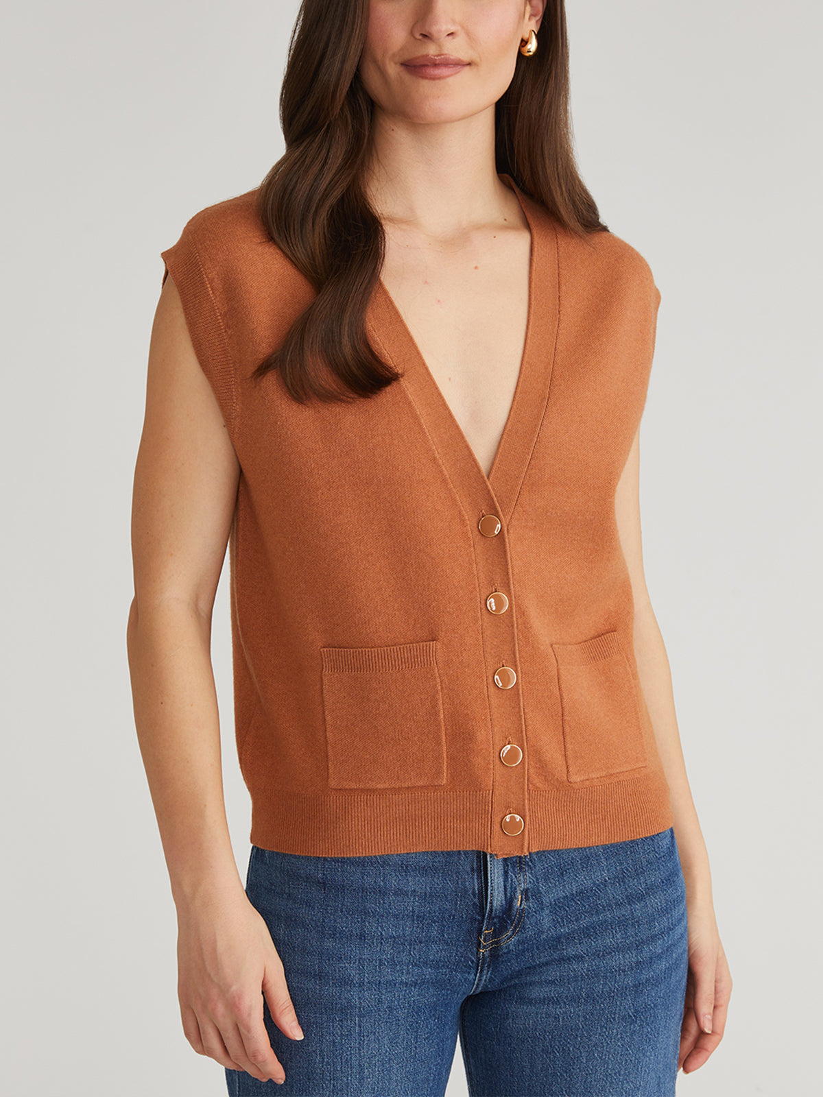 Wearing a stylish 525 America sleeveless brown button-down cardigan layered over blue jeans, featuring two pockets, they stand against a plain background. Their long, wavy hair enhances the look as they maintain a calm expression without smiling.