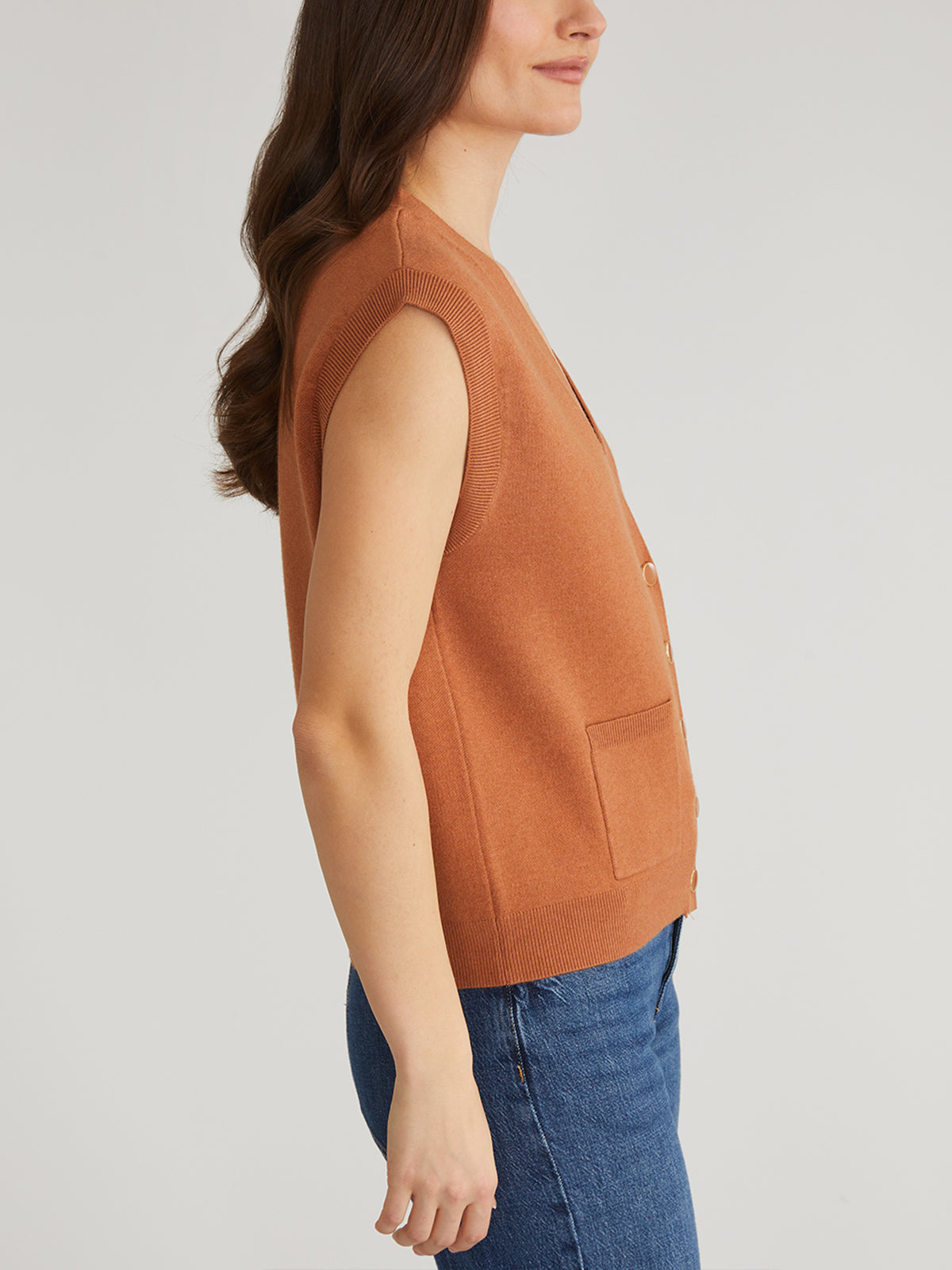 A person with long brown hair stands sideways, wearing a 525 America button-down sleeveless brown sweater vest with front pockets over blue jeans on a plain gray background; the vest is neatly buttoned.