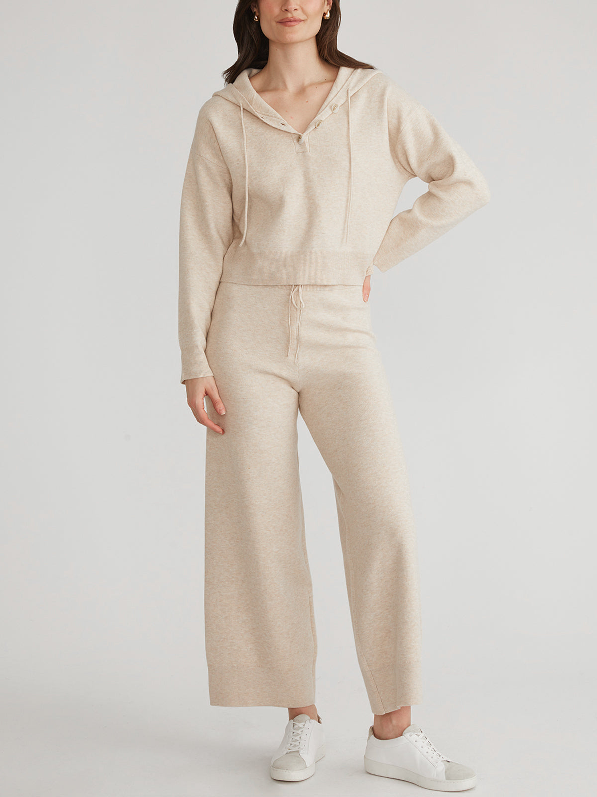 Wearing a beige 525 America hooded sweater with matching wide-legged pants, a person stands against a plain background. The relaxed fit is enhanced by white sneakers, and one hand rests on their hip.