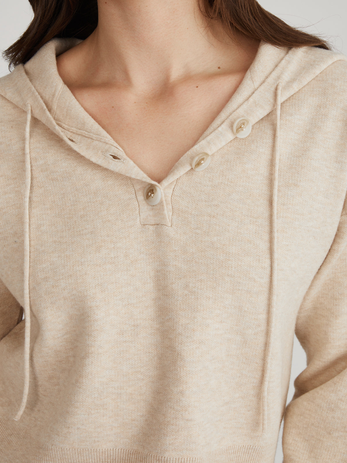 A close-up shows a person in 525 Americas beige relaxed-fit hooded sweater with a V-neck and three buttons, highlighting its cozy hood and drawstring. The persons head is not visible.