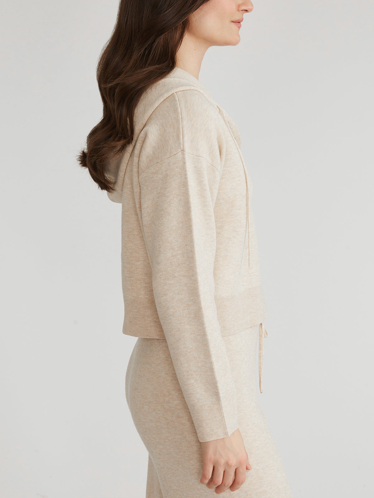 A woman in a 525 America light beige knit sweater with a cozy hood and matching pants stands in profile. Her long brown hair, styled in loose waves, cascades down her back, complementing the relaxed fit vibe against the neutral white background.