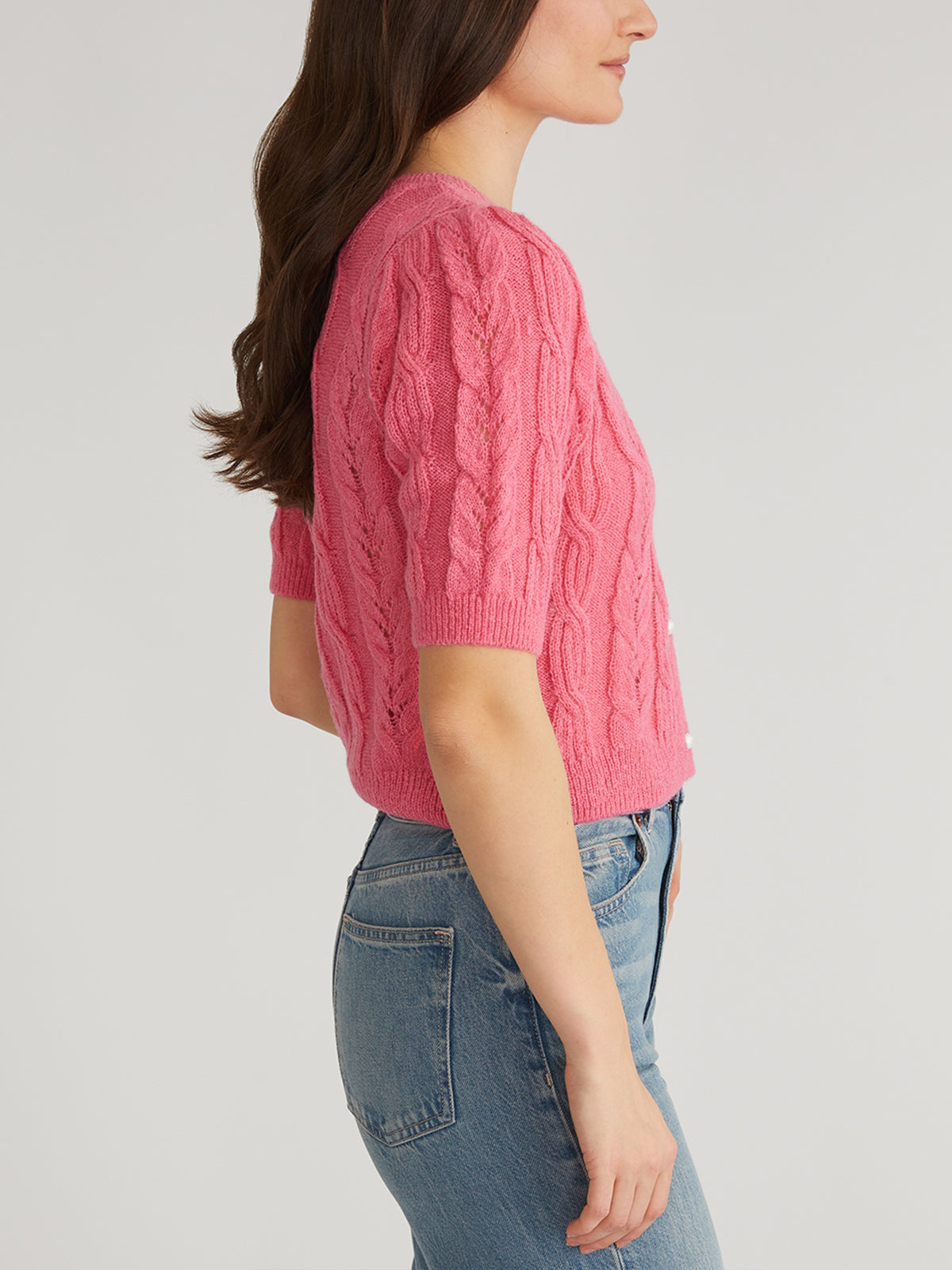A person with long brown hair wears a pink cable-knit short-sleeve sweater from 525 America, featuring pearl-like buttons, paired with blue jeans. They stand sideways, facing left against a plain, light-colored background.