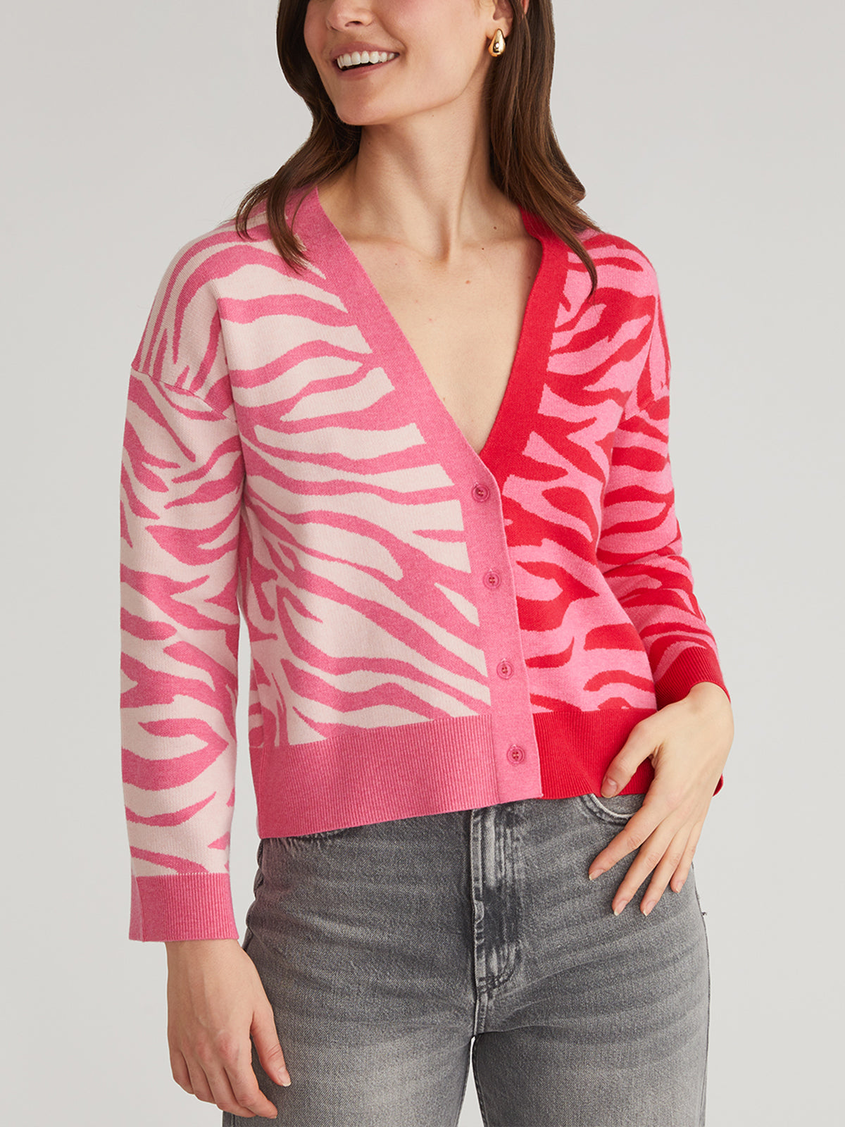 A person wearing a 525 America relaxed fit Colorblock Zebra Print Cardigan in pink and red, featuring a deep V-neck and button closure. The cardigan has different shades on each side. They pair it with gray jeans and have long hair.