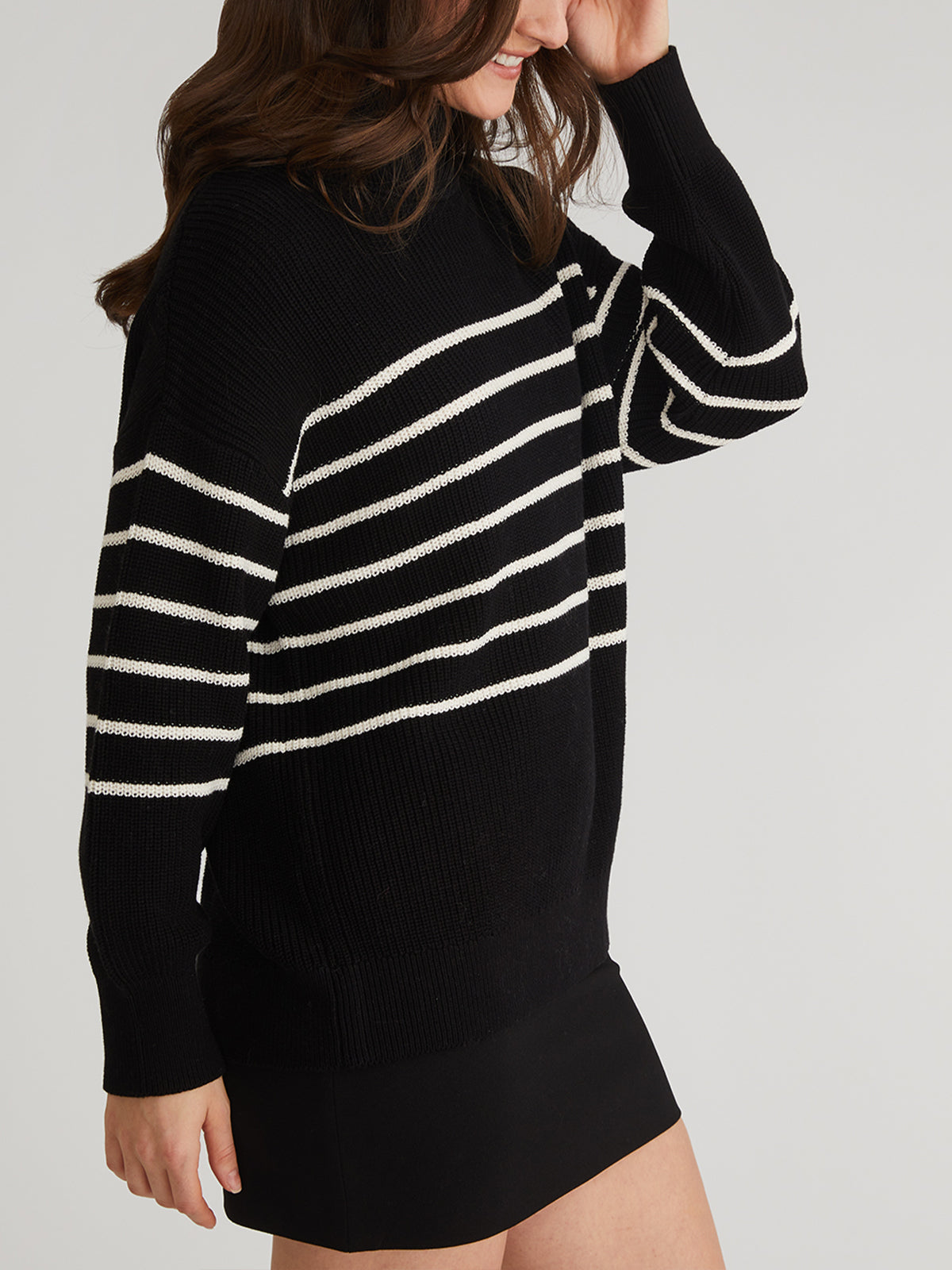 A person dons a 525 America oversize turtleneck sweater, featuring white horizontal stripes on 100% cotton, paired with a black skirt. The light backdrop highlights their style as they softly touch their hair, with their face partially out of frame.