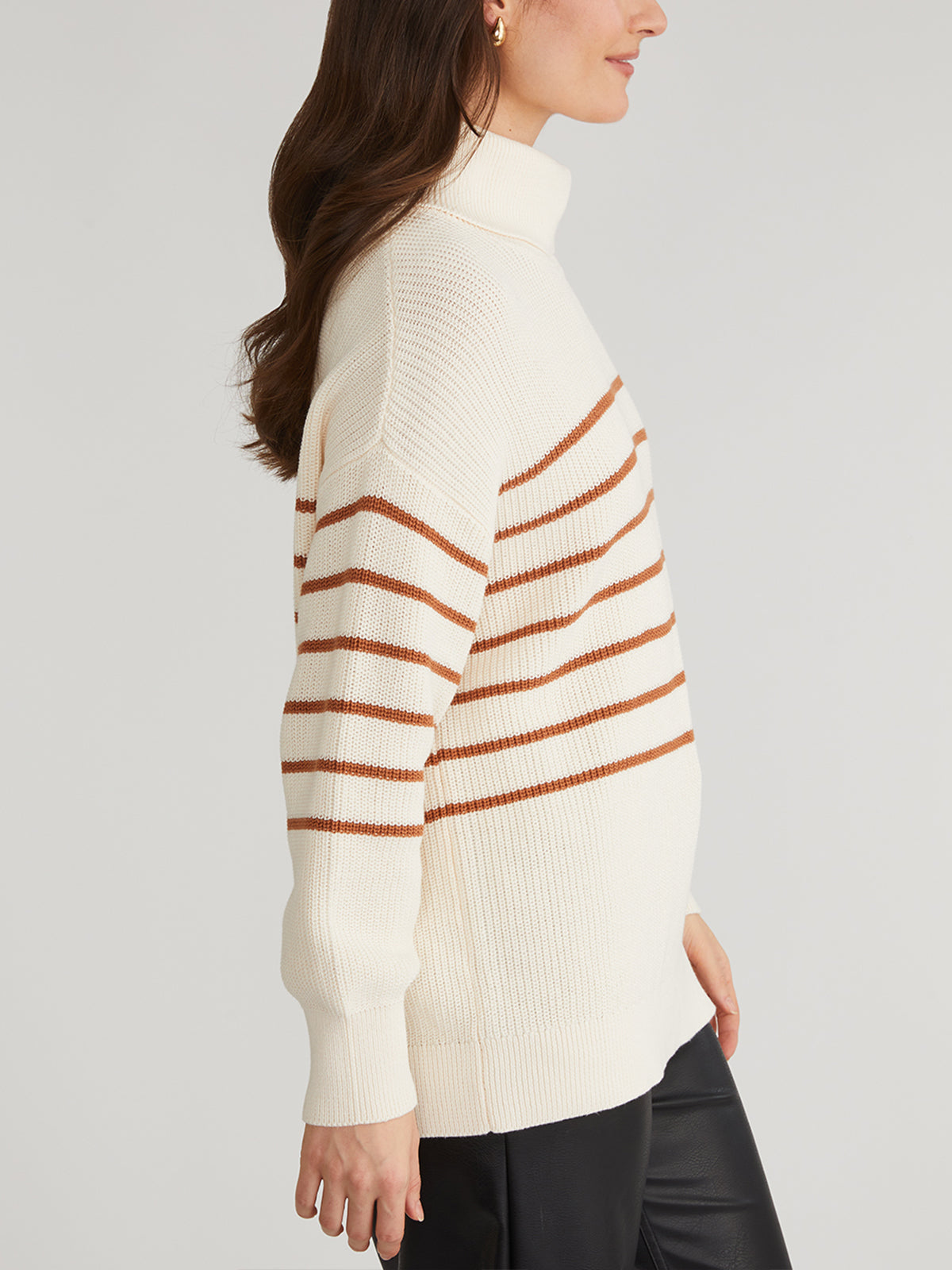 The individual is wearing an oversize stripe turtleneck from 525 America, a cream sweater with brown stripes. The long sleeves pair well with black pants against a plain, light backdrop. Made of 100% cotton, it provides style and comfort.