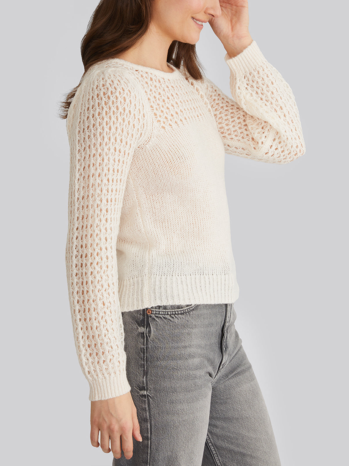 A person with long brown hair, wearing a 525 America white knitted sweater with breathable mesh sleeves and light gray jeans, faces sideways while touching their head against a plain light gray backdrop.