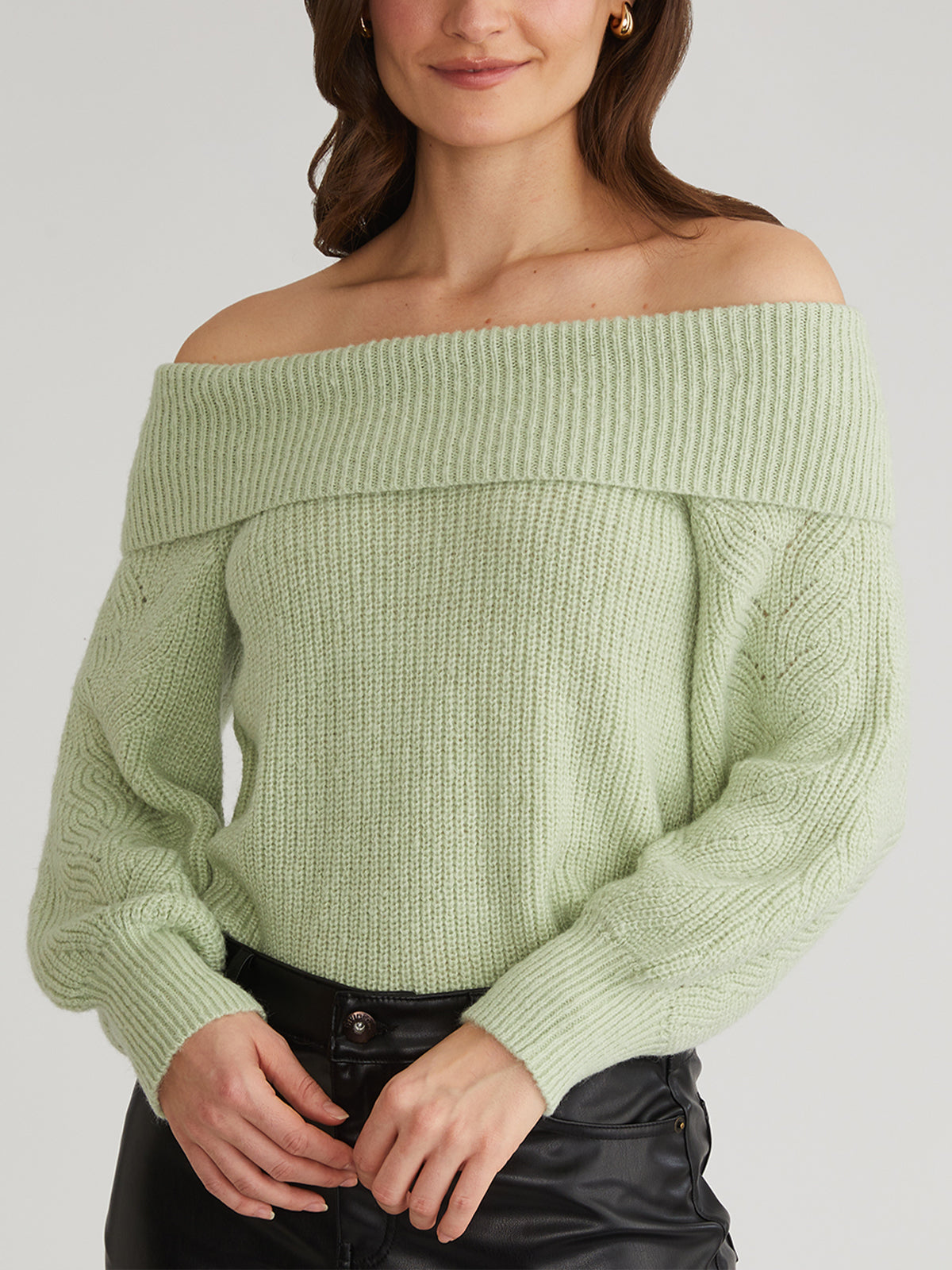 A person sports an off-the-shoulder, light green knitted 525 America sweater with black pants. This relaxed pullover boasts a large folded-over collar and long sleeves. Their hands are lightly clasped in front against a plain gray background, ideal for casual outings.
