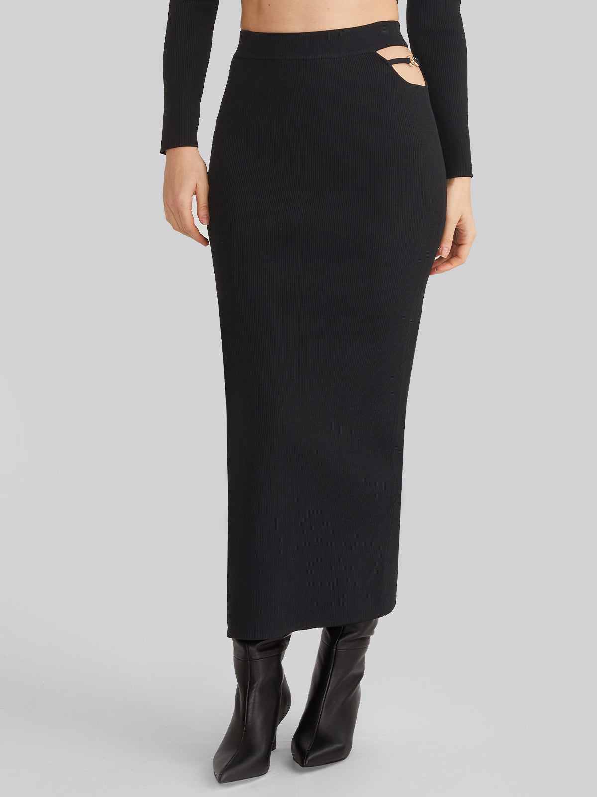 A person wears a long, form-fitting black maxi skirt with side cutouts and a matching long-sleeve top by 525 America. The outfit includes black heeled boots with buckled straps against a plain gray background.