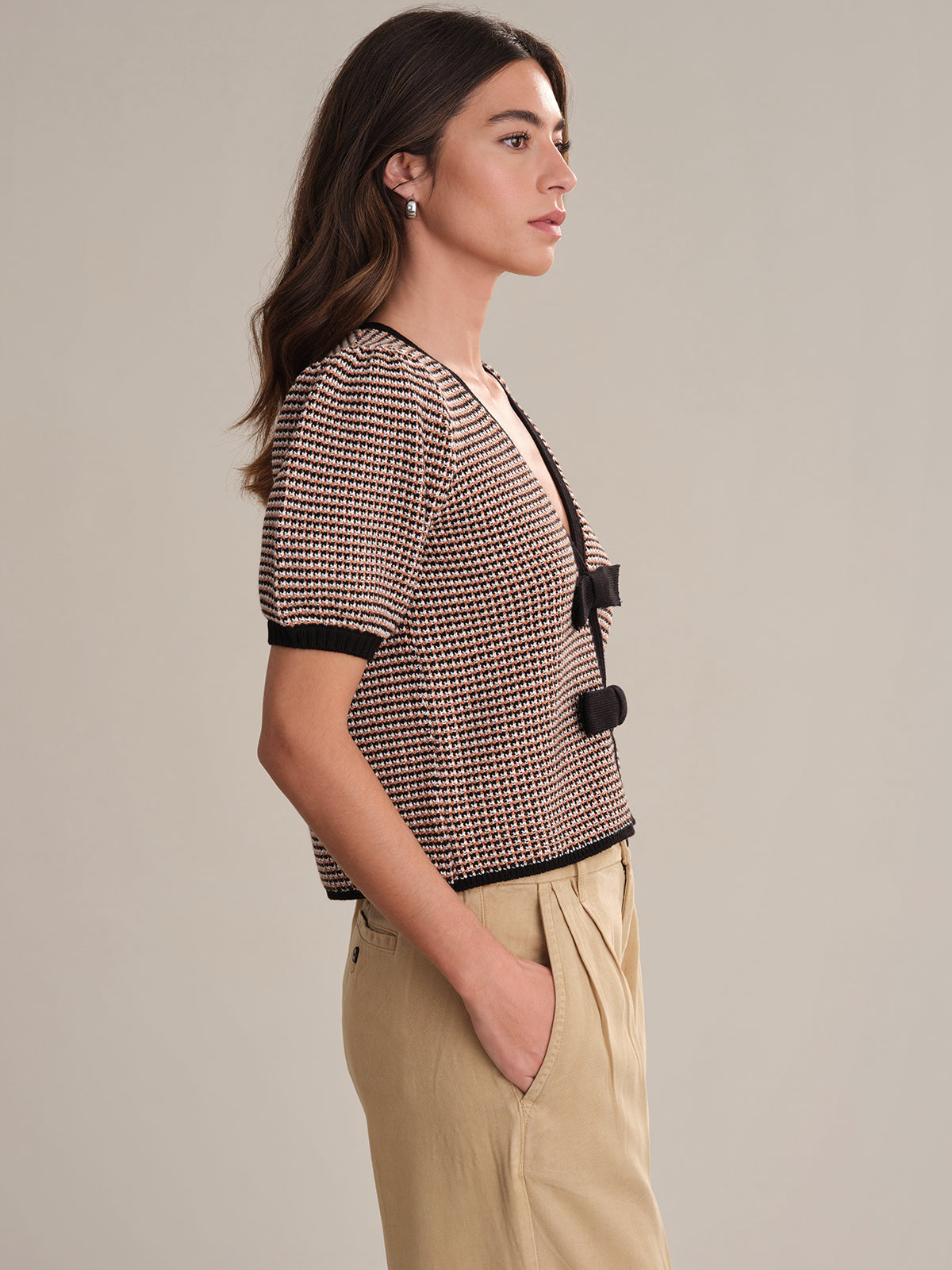 A woman in profile wears beige pants with her hands in the pockets and a 525 America patterned knit top featuring black trim and bow ties, inspired by a feminine cardigan. The background is plain and neutral.