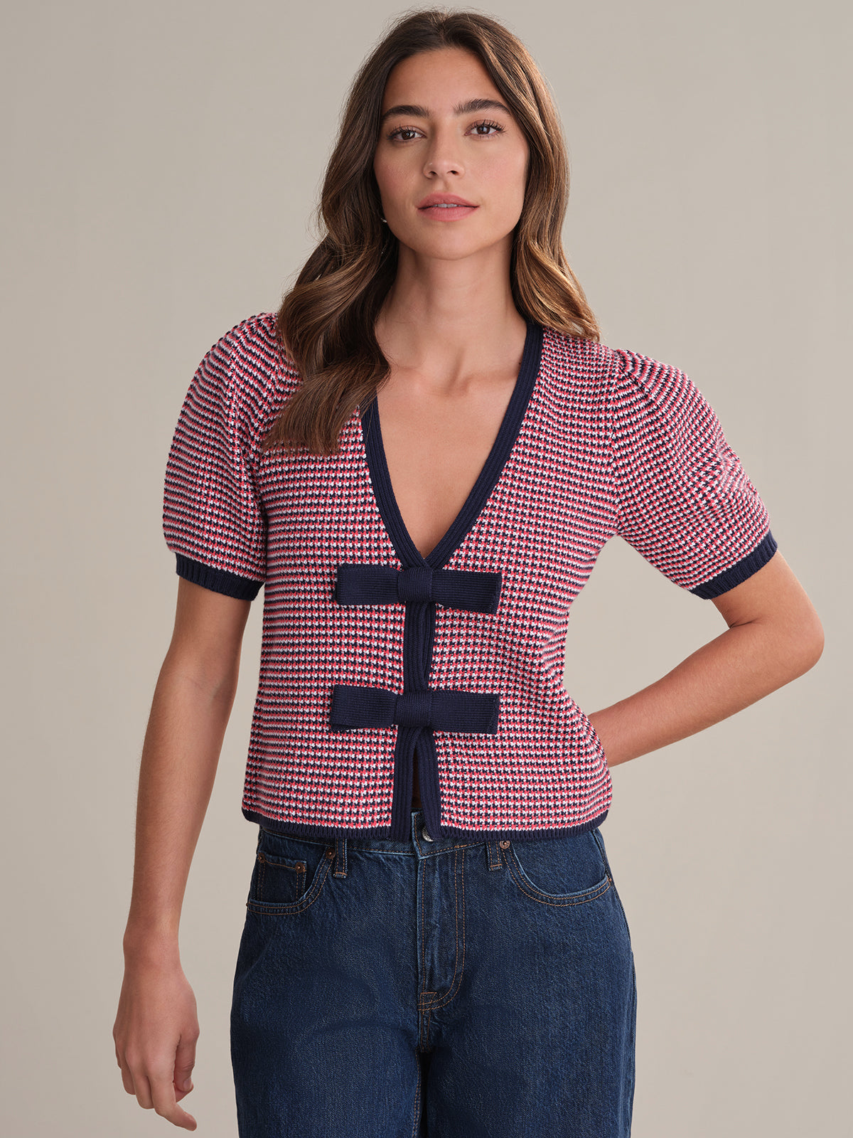 A woman faces forward in a 525 America short-sleeve cardigan with a red and white houndstooth pattern, navy trim, two horizontal navy bow ties, and high-waisted blue jeans. The background is plain.