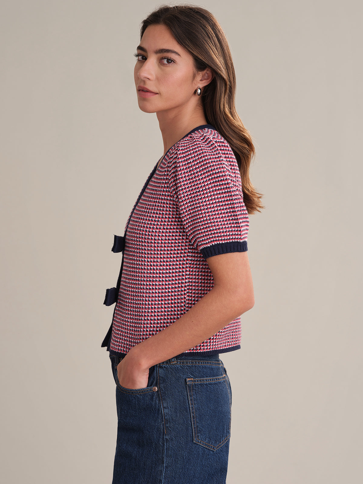 A woman with long brown hair stands in profile, wearing a 525 America feminine red and navy short-sleeved cardigan with two front bows and high-waisted blue jeans. One hand is in her pocket as she faces left.