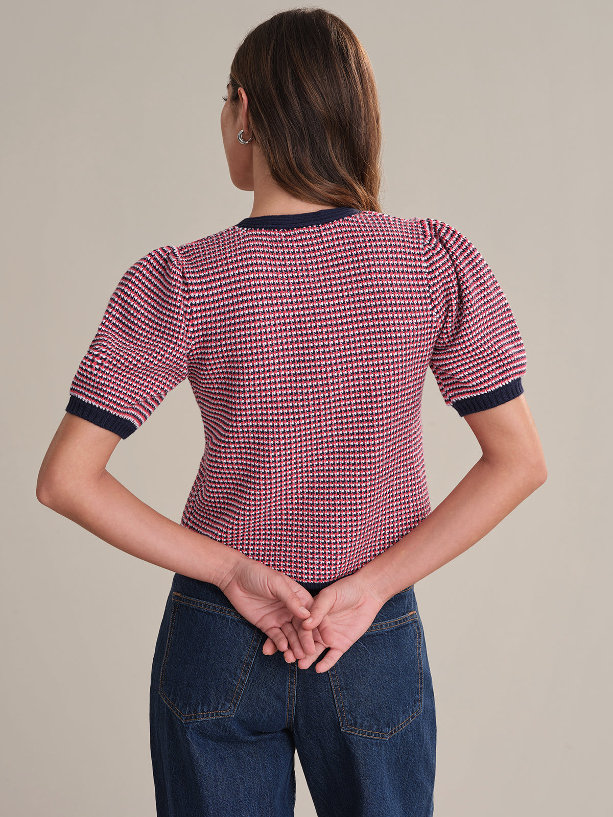 A person faces away from the camera with hands behind their back, wearing a 525 America red and blue checkered knit top with puff sleeves and blue jeans against a plain background.
