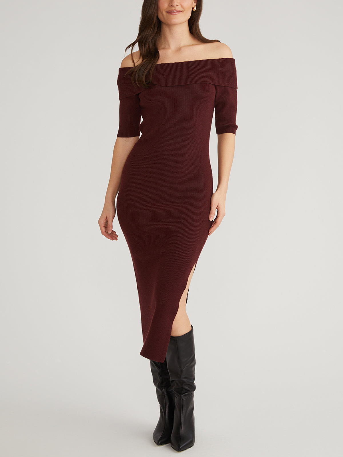 A woman is wearing a maroon ribbed midi dress from 525 America, with an elegant off-shoulder neckline, sleek silhouette, elbow-length sleeves, and a subtle side slit. Her long brown hair complements her black knee-high boots against a light gray background.