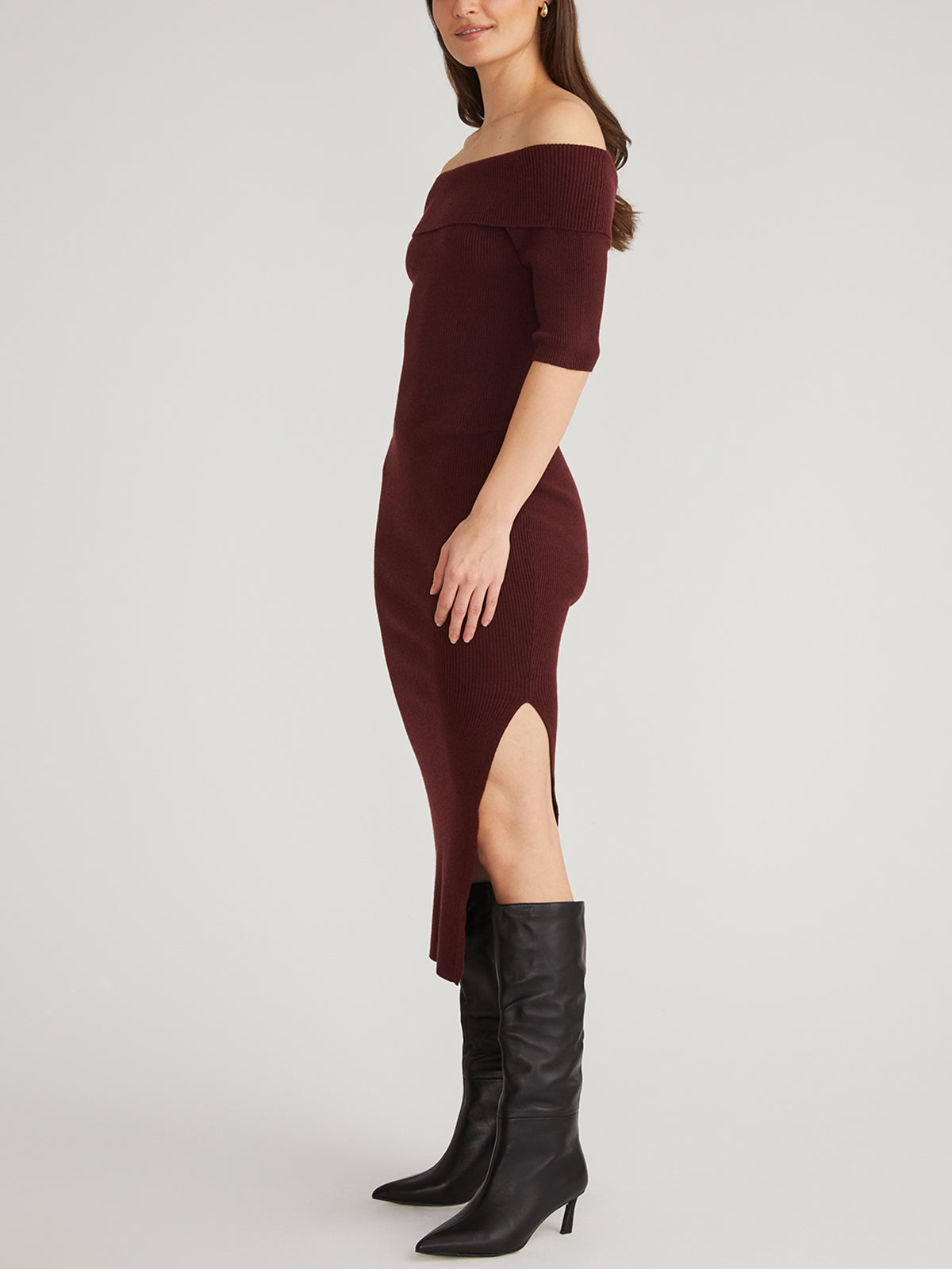 A person stands confidently against a plain background wearing an off-shoulder, burgundy ribbed midi dress with three-quarter sleeves and a side slit by 525 America, paired elegantly with black knee-high boots.