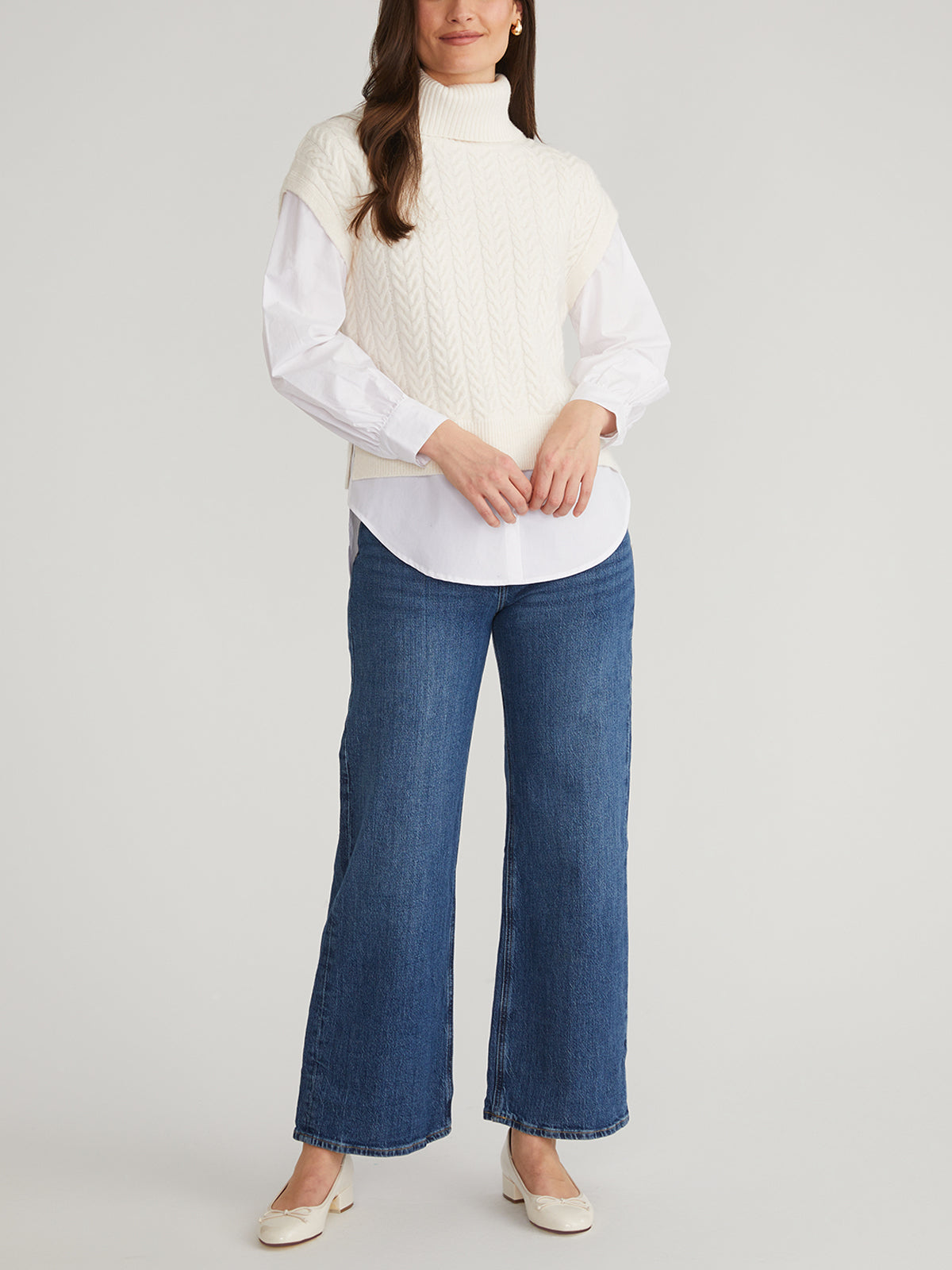 A person stands against a plain background wearing a 525 America turtleneck sweater, a white long-sleeve shirt, blue wide-leg jeans, and beige heels. Their arms are relaxed by their sides.
