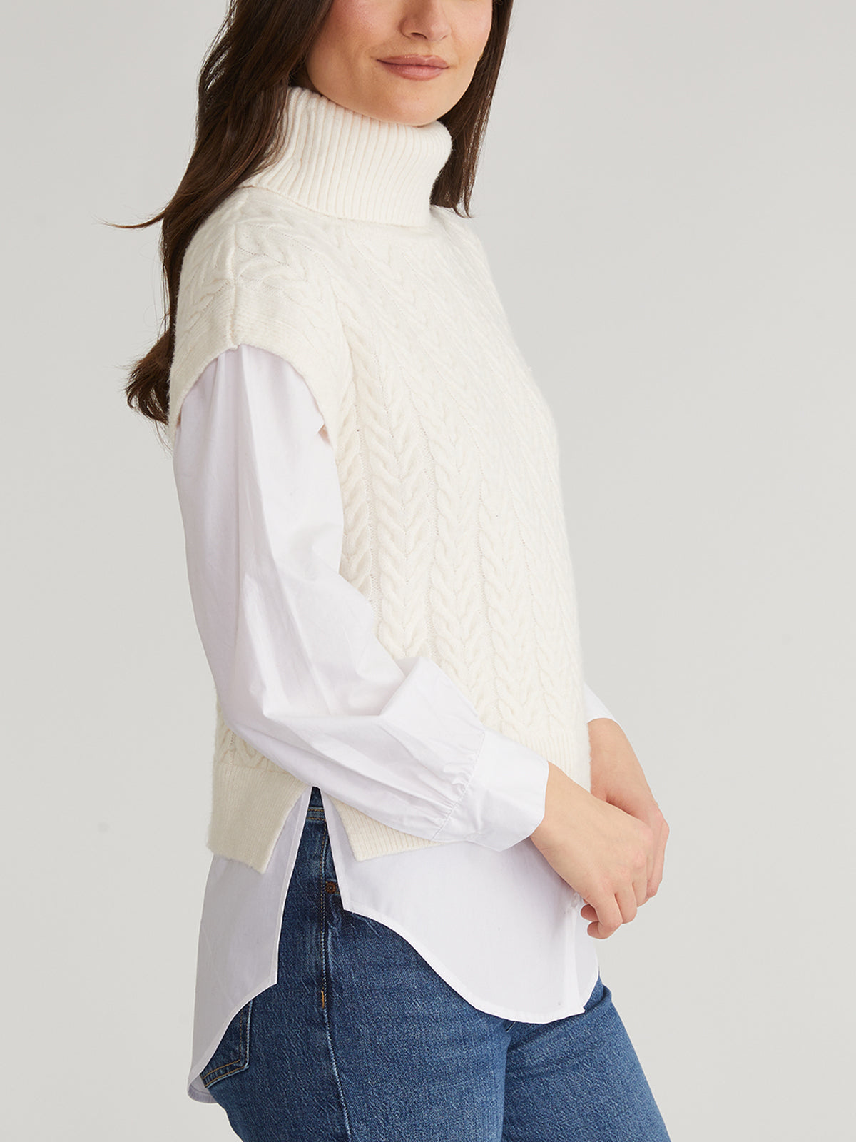 A person wearing a cream-colored Sofia turtleneck sweater by 525 America is layered over a white long-sleeve shirt with denim jeans, standing sideways against a plain light background.