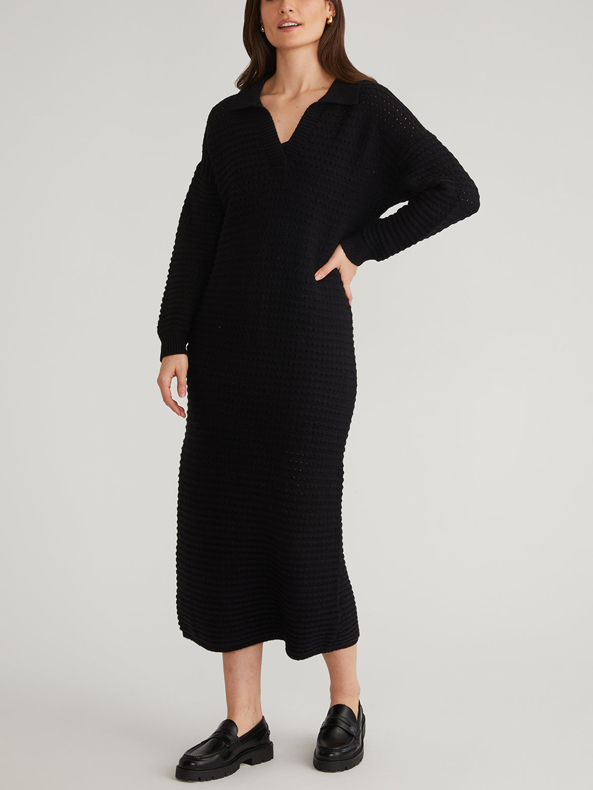 A person in a 525 America Raya Polo Dress, known for its heavyweight stretch knit and pointelle stitching, stands confidently with one hand on their hip. The long-sleeved dress has a V-neckline and is matched with black slip-on shoes against a plain, light background.