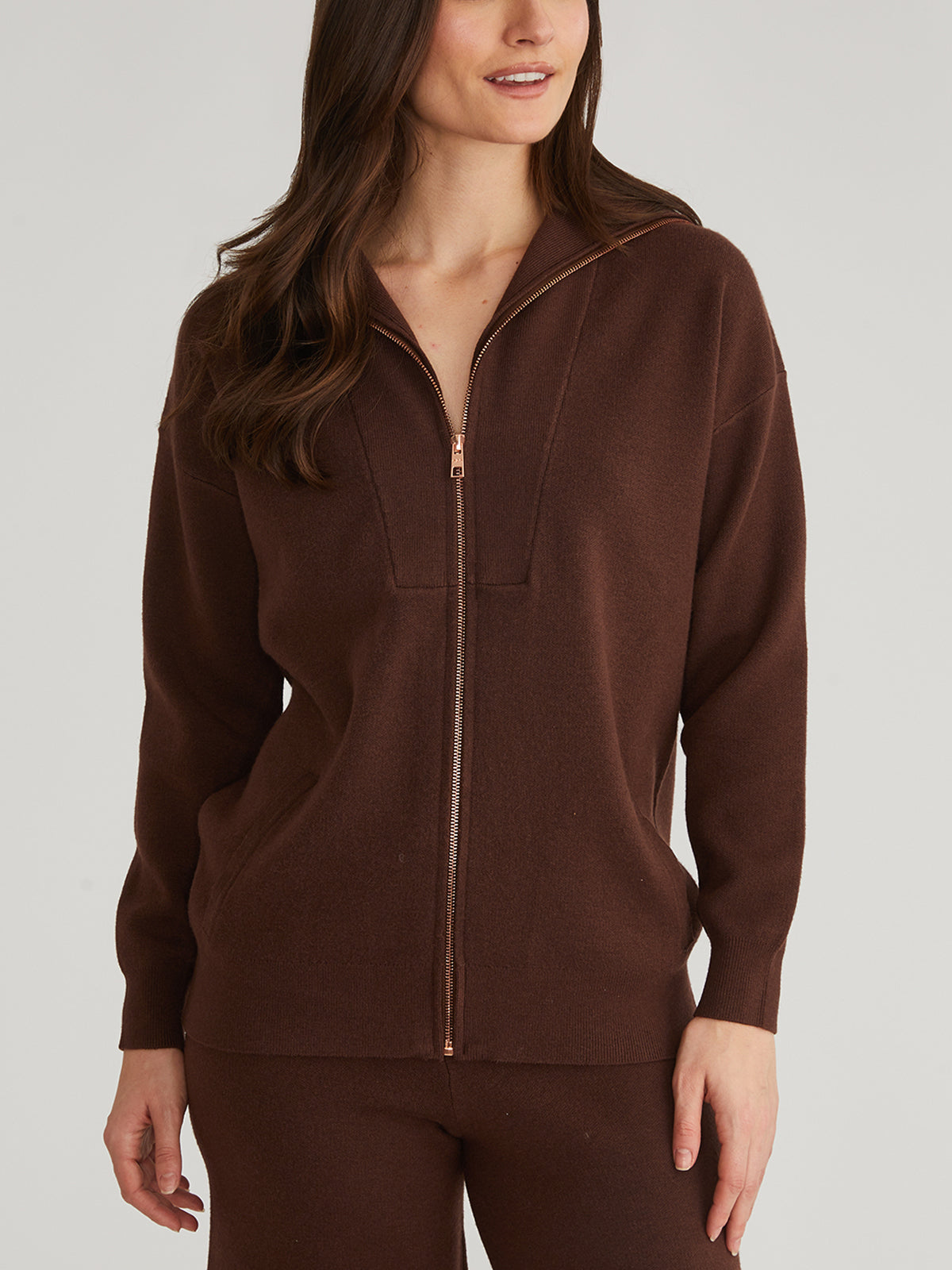 They sport an oversized brown zip-up sweater from 525 America, made with luxe yarn, featuring a front zipper and relaxed collar, paired with matching pants. Their long brown hair cascades down as their right hand rests casually by their side against a plain gray background.