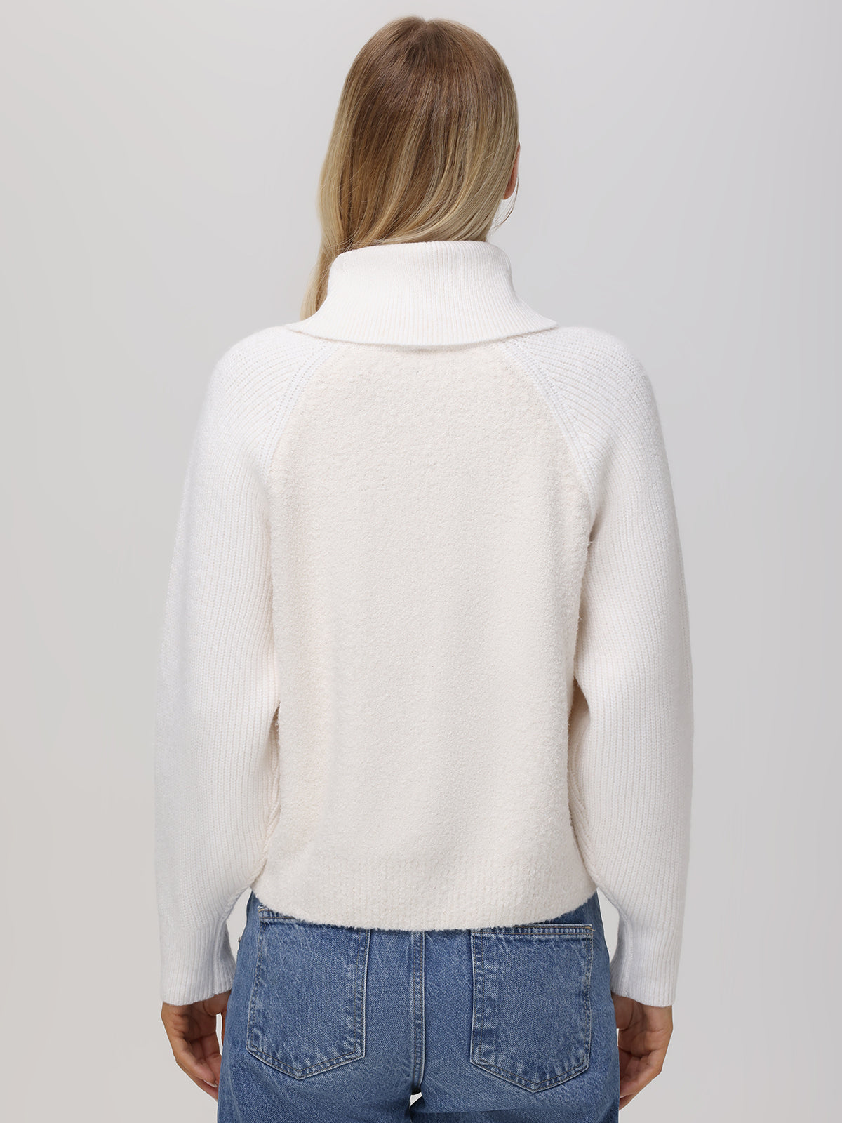 A person with long hair, seen from the back, wears a white textured turtleneck sweater by 525 America with raglan sleeves and blue jeans. The sweaters ribbed design is highlighted against a plain light gray background.