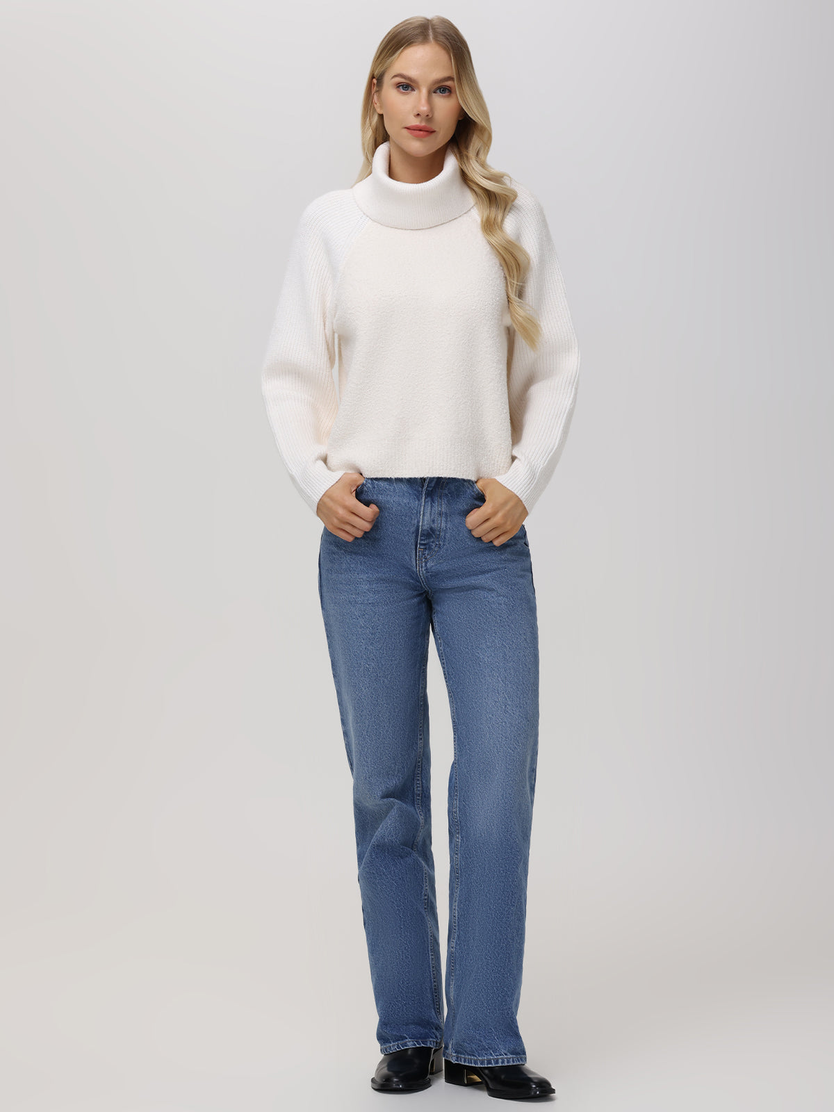 A person stands against a plain background wearing a 525 America white turtleneck sweater with raglan sleeves, paired with blue jeans. They sport long blonde hair and black shoes, with hands casually in their pockets.