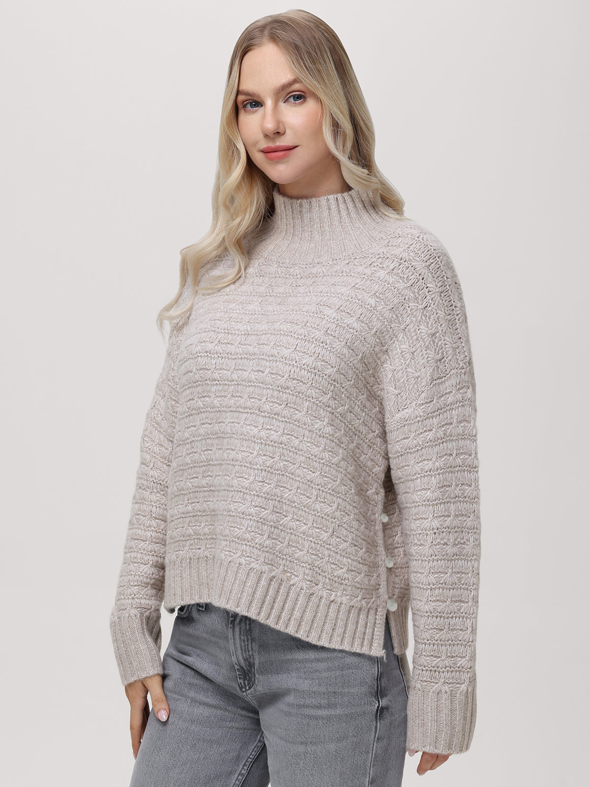 A woman with long blonde hair wears a 525 America light gray textured knit mockneck pullover and light blue jeans, standing against a plain light background and looking at the camera with a faint smile.