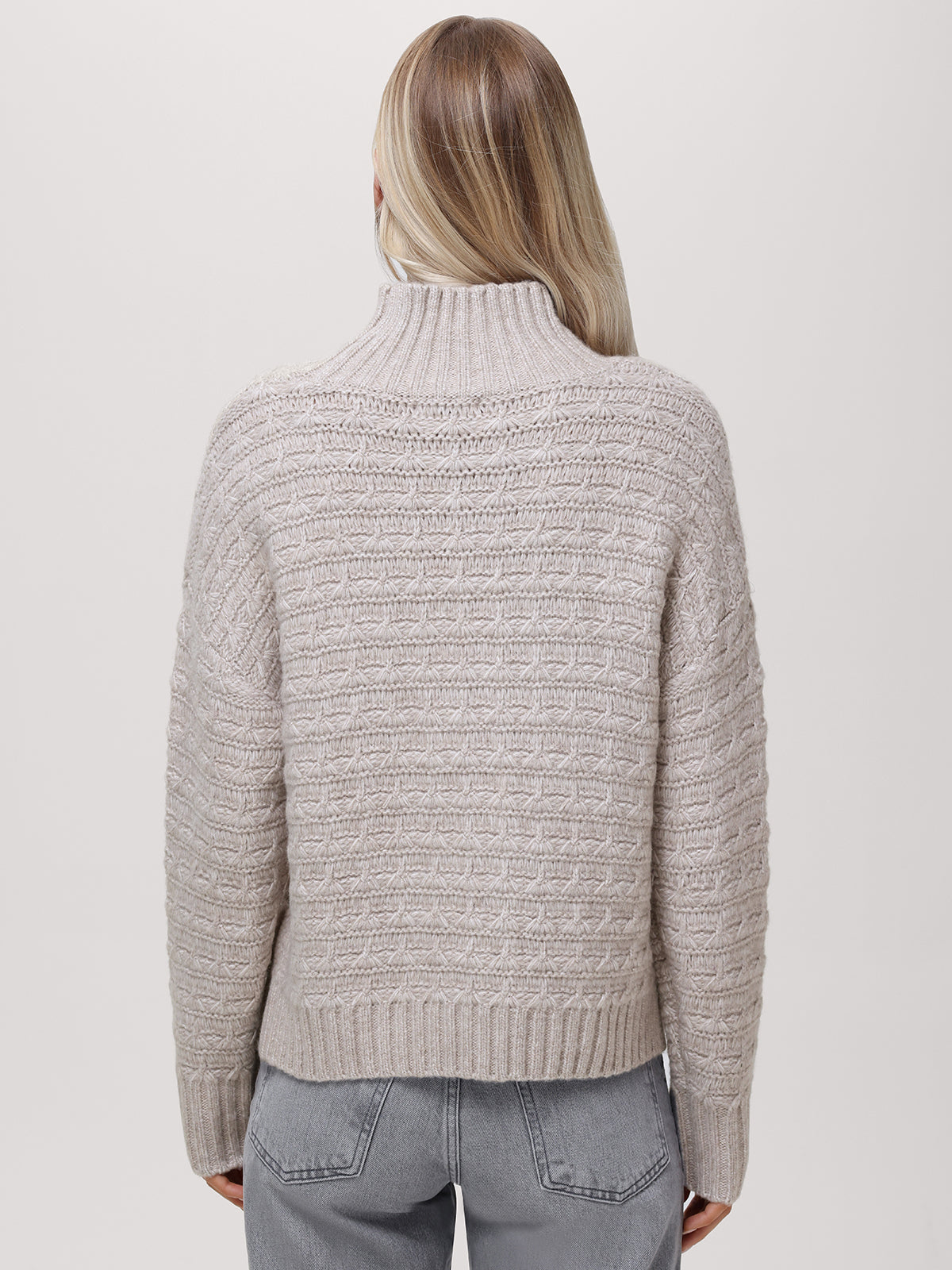 A person with long, straight blonde hair is shown from the back, wearing a relaxed fit, textured light beige mockneck pullover sweater by 525 America and light gray jeans against a plain light background.