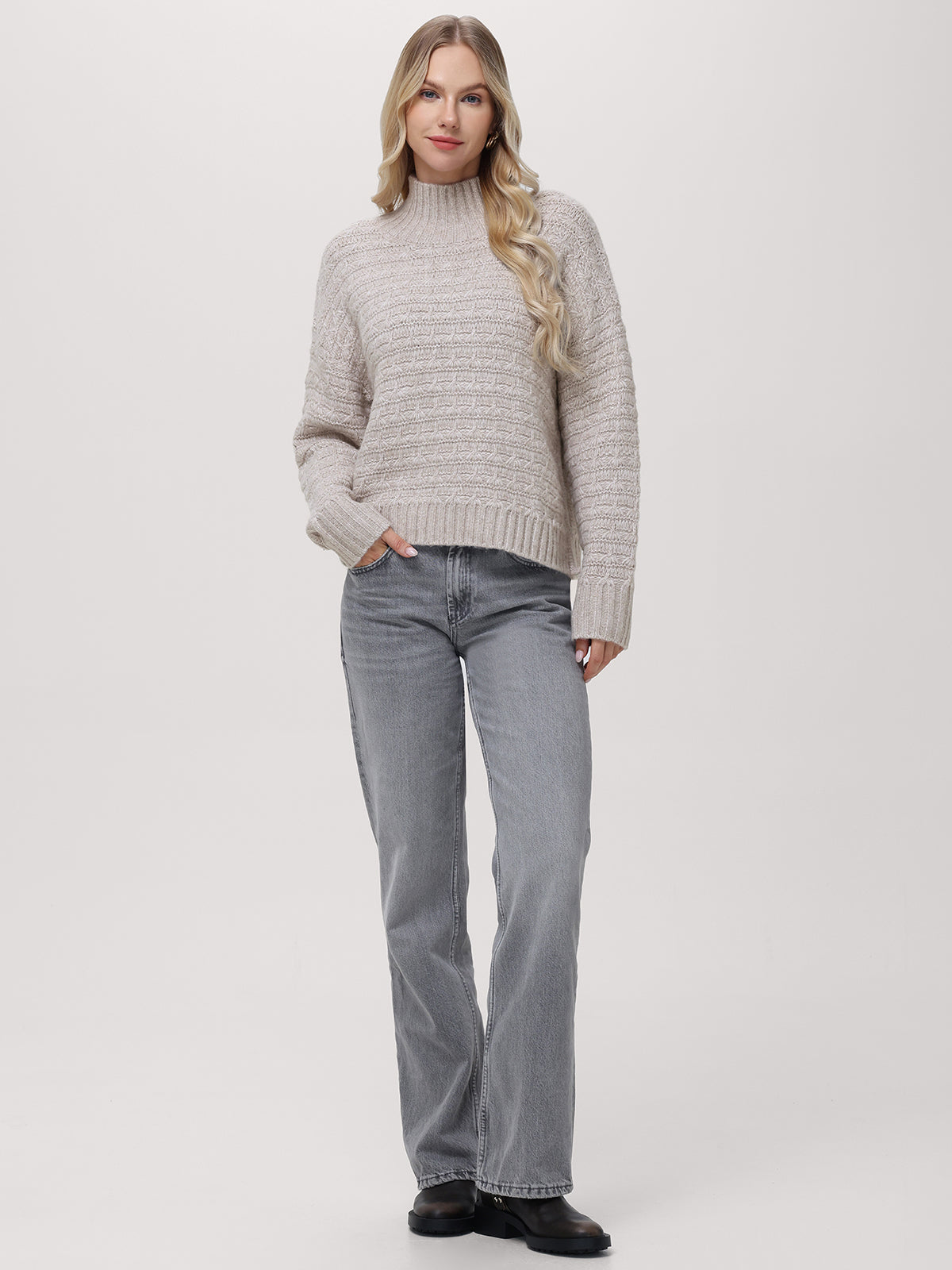 A woman with long, wavy blonde hair stands against a plain background wearing a relaxed fit, light gray mockneck pullover sweater with subtle buttons by 525 America, paired with matching jeans and black shoes, one hand in her pocket.