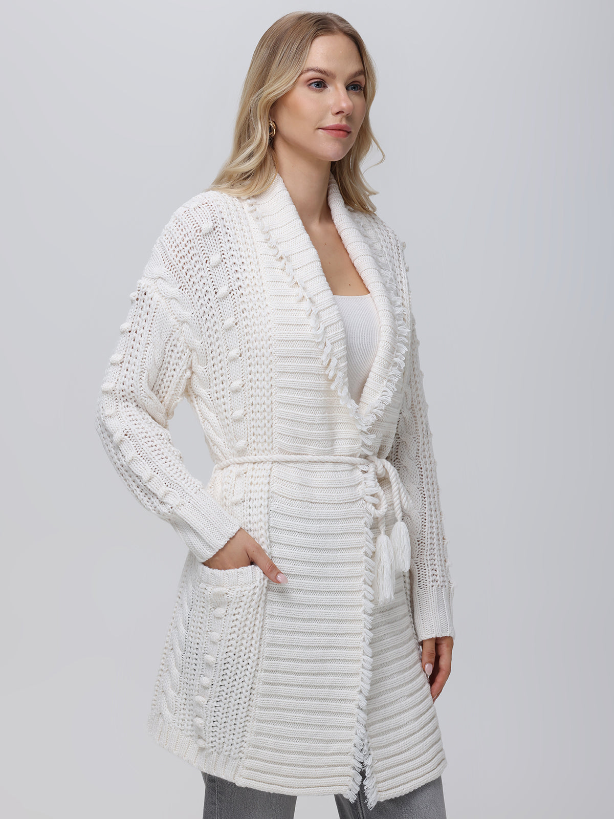 A woman exudes stylish sophistication in a 525 America Penelope Cable Sweater Coat, featuring a textured knit with pockets and a tie belt. Against a plain backdrop, she pairs it with long blonde hair, a white top, and light-colored pants.