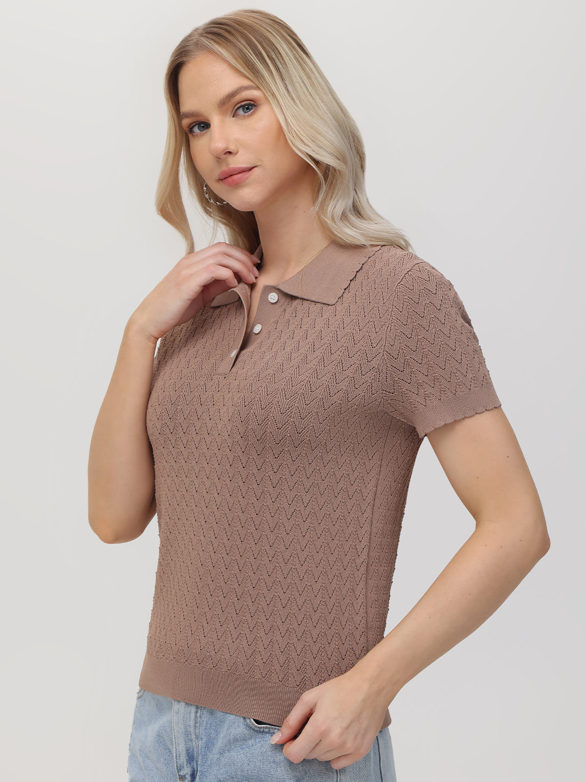 A woman with long blonde hair models a 525 America light brown short-sleeve knit classic polo, highlighting a zigzag pattern, paired with light blue jeans. She stands against a plain backdrop, her right hand lightly touching her hair while gazing at the camera.