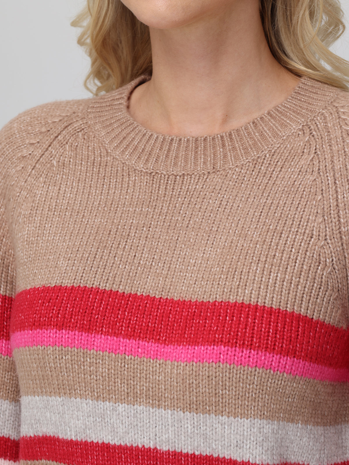 A woman wears a 525 America knitted cropped pullover with colorblock stripes in brown, red, pink, and white. The fabrics texture and raglan sleeves are highlighted. A plain neutral backdrop frames her partially obscured face.