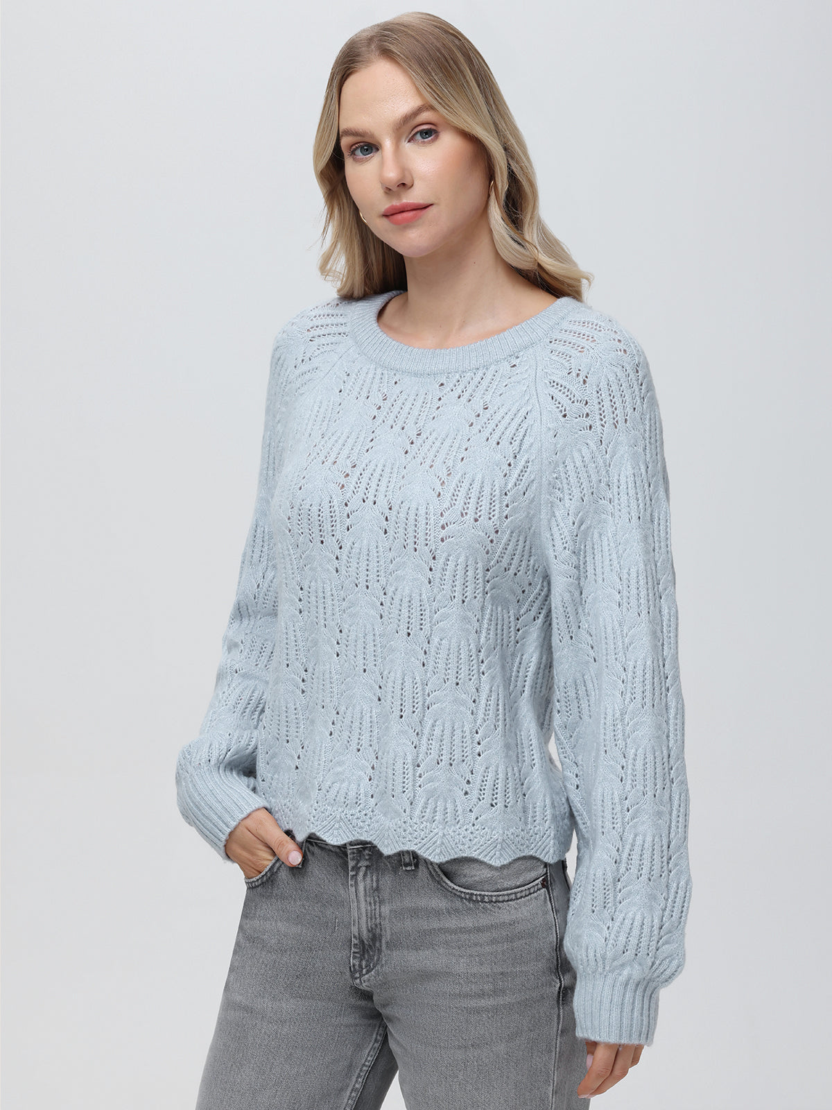 Against a plain backdrop, a person wears a light blue 525 America knitted pullover featuring a crew neckline and patterned design, paired with gray jeans. Their hair is down, one hand rests in their pocket as they face the camera with a neutral expression.