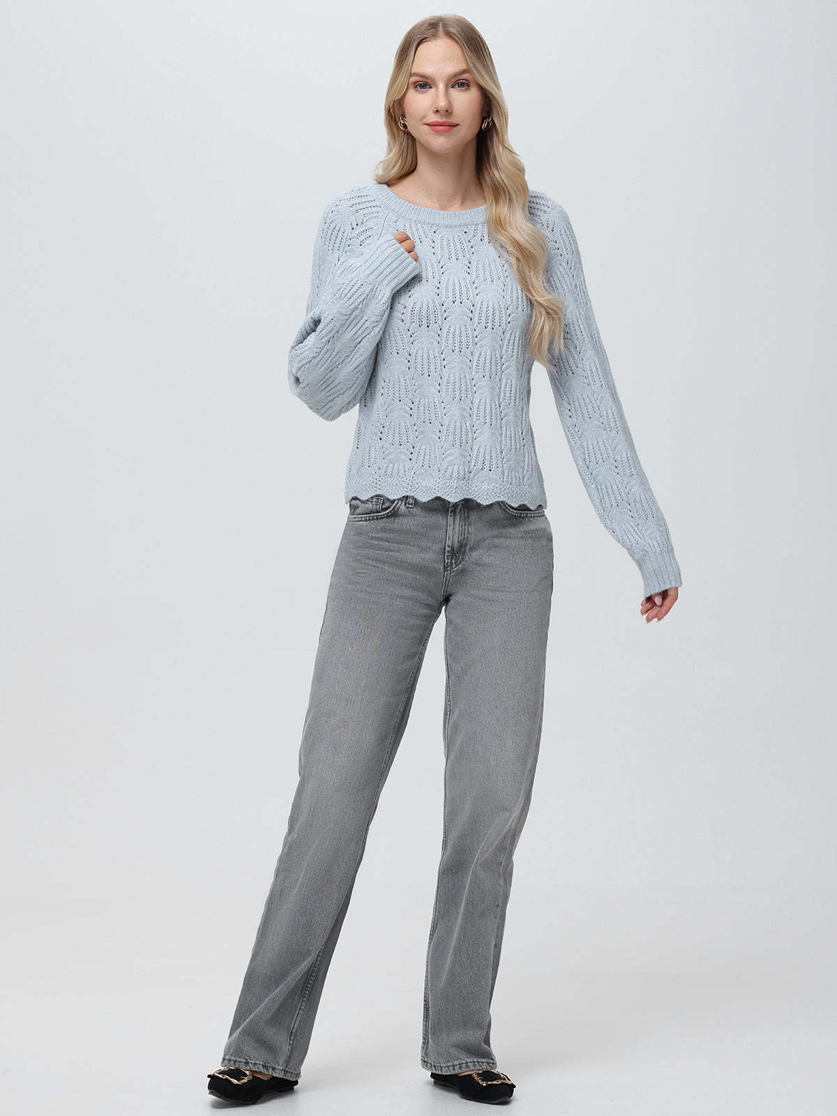 A person with long blonde hair is wearing a 525 America light blue knit pullover with a crew neckline and gray jeans, posed against a plain background, looking forward. One hand rests on the sweater while the other hangs by their side.