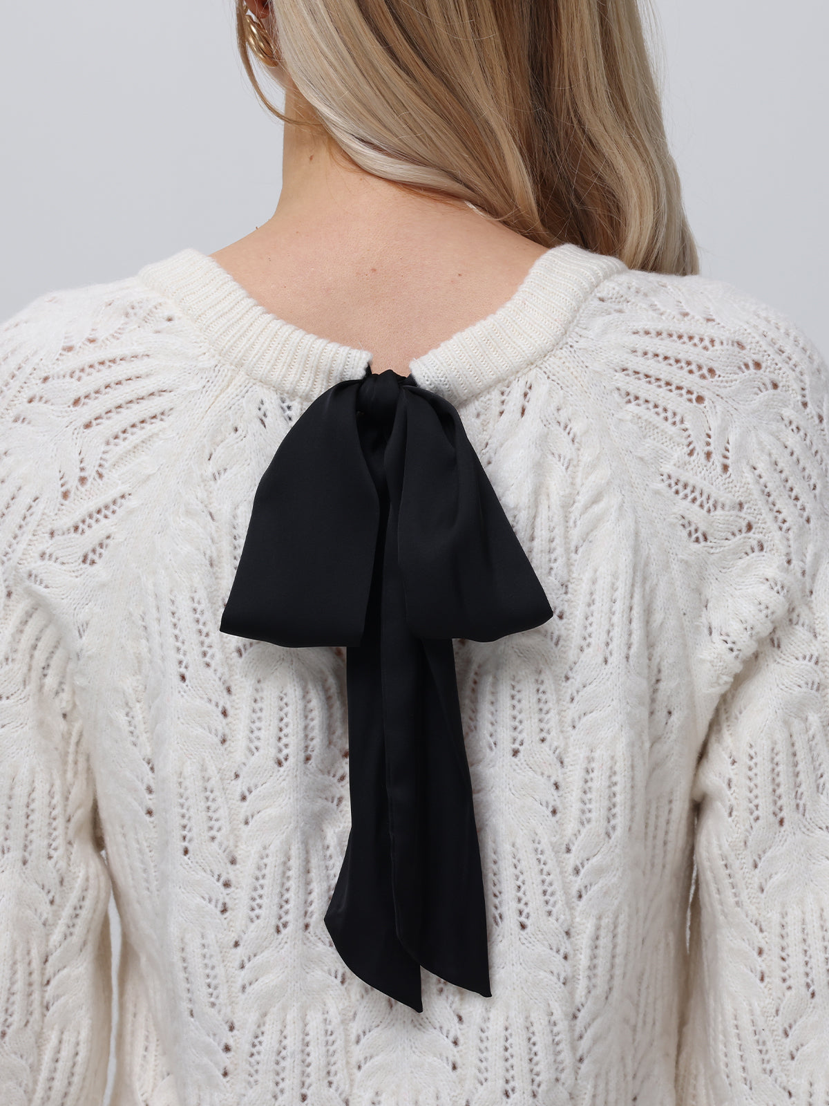 A person in a 525 America cream pullover sweater with a large black bow and pointelle detailing is seen from behind. Long ribbons drape down the back, and their light-colored hair falls over one shoulder.