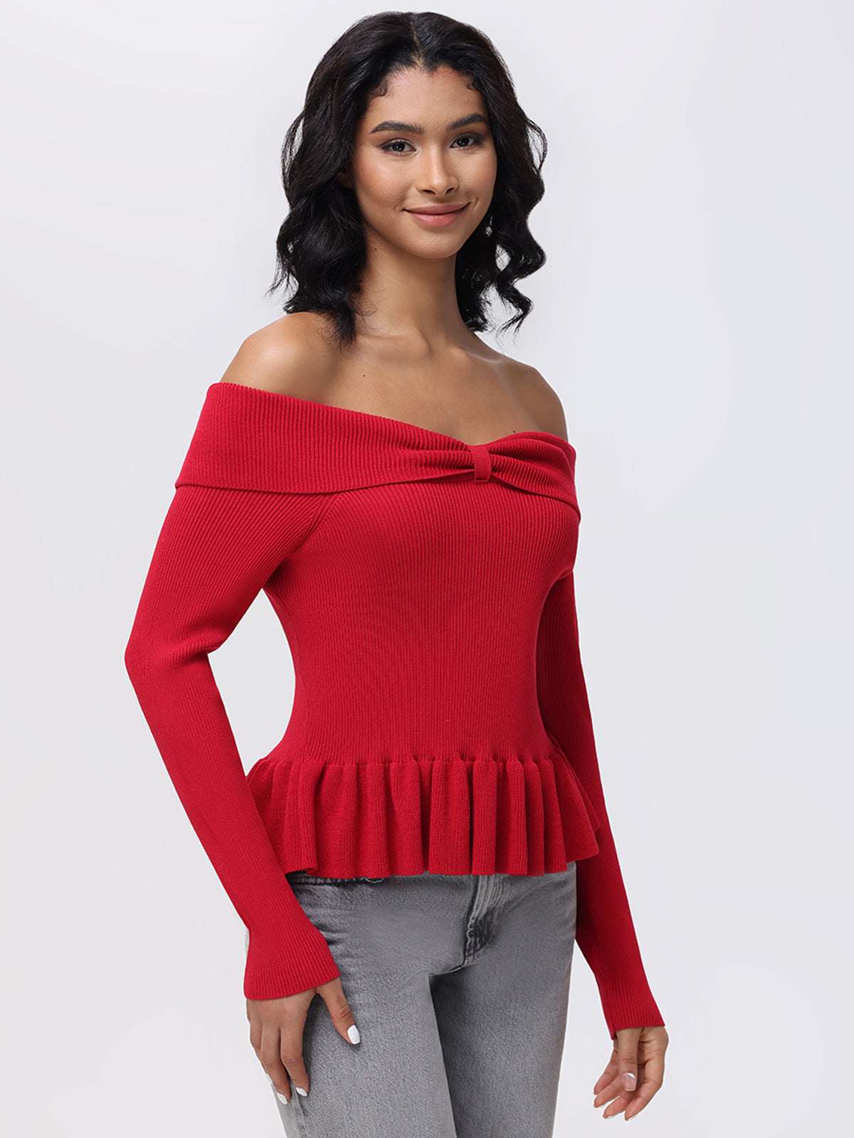 A person with long black hair wears a red 525 America Off Shoulder Peplum Pullover, featuring chic ruffles at the waist and a bow detail, paired with gray jeans. They stand against a plain light background, effortlessly embodying stylish elegance.