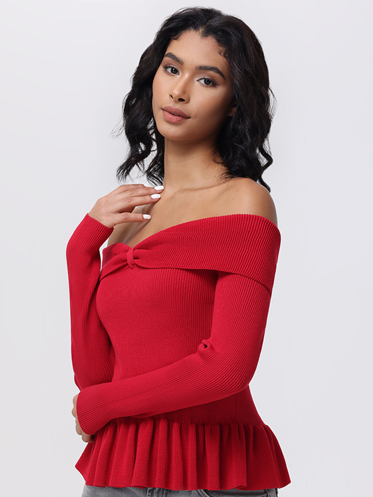 A person with shoulder-length curly hair wears the chic Morgan Off Shoulder Peplum Pullover by 525 America. This red top features long sleeves and a pleated hem, adding elegance as they stand against a plain background, gazing slightly to the side.