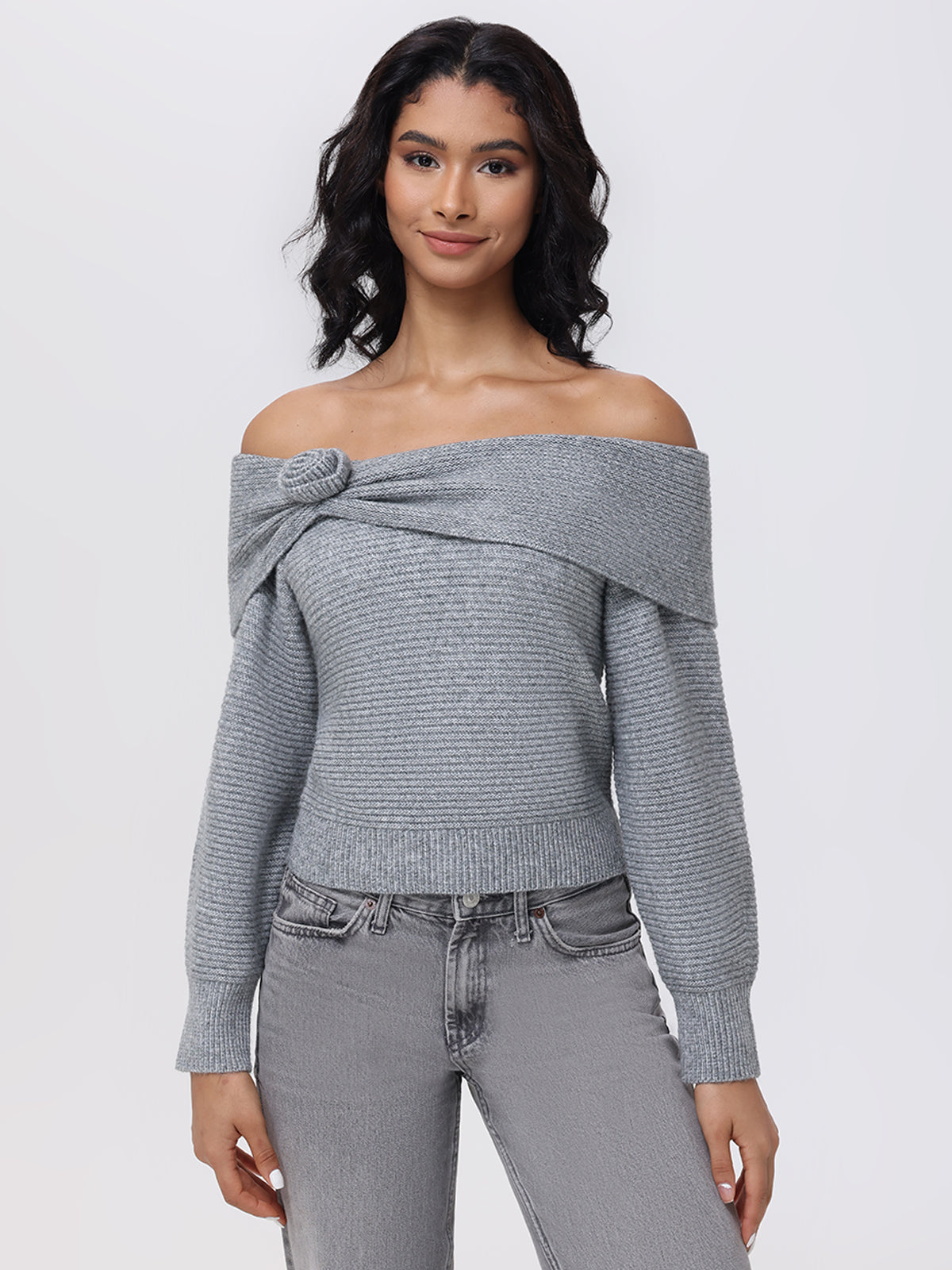 With long, wavy hair, a person wears the 525 America Elizabeth Sweater, light gray and off-the-shoulder with a knot detail, paired with gray jeans. Set against a plain white background, they gaze directly at the camera with a neutral expression.