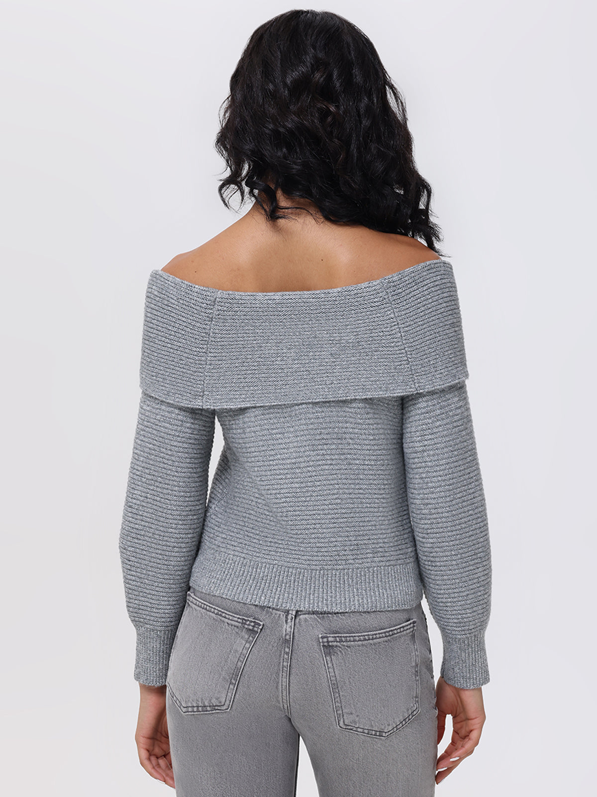 The individual, turned away from the camera, models a chic off-the-shoulder design by 525 America in an Elizabeth Sweater, paired with gray jeans. The scene is minimalist, highlighted by a simple white backdrop.