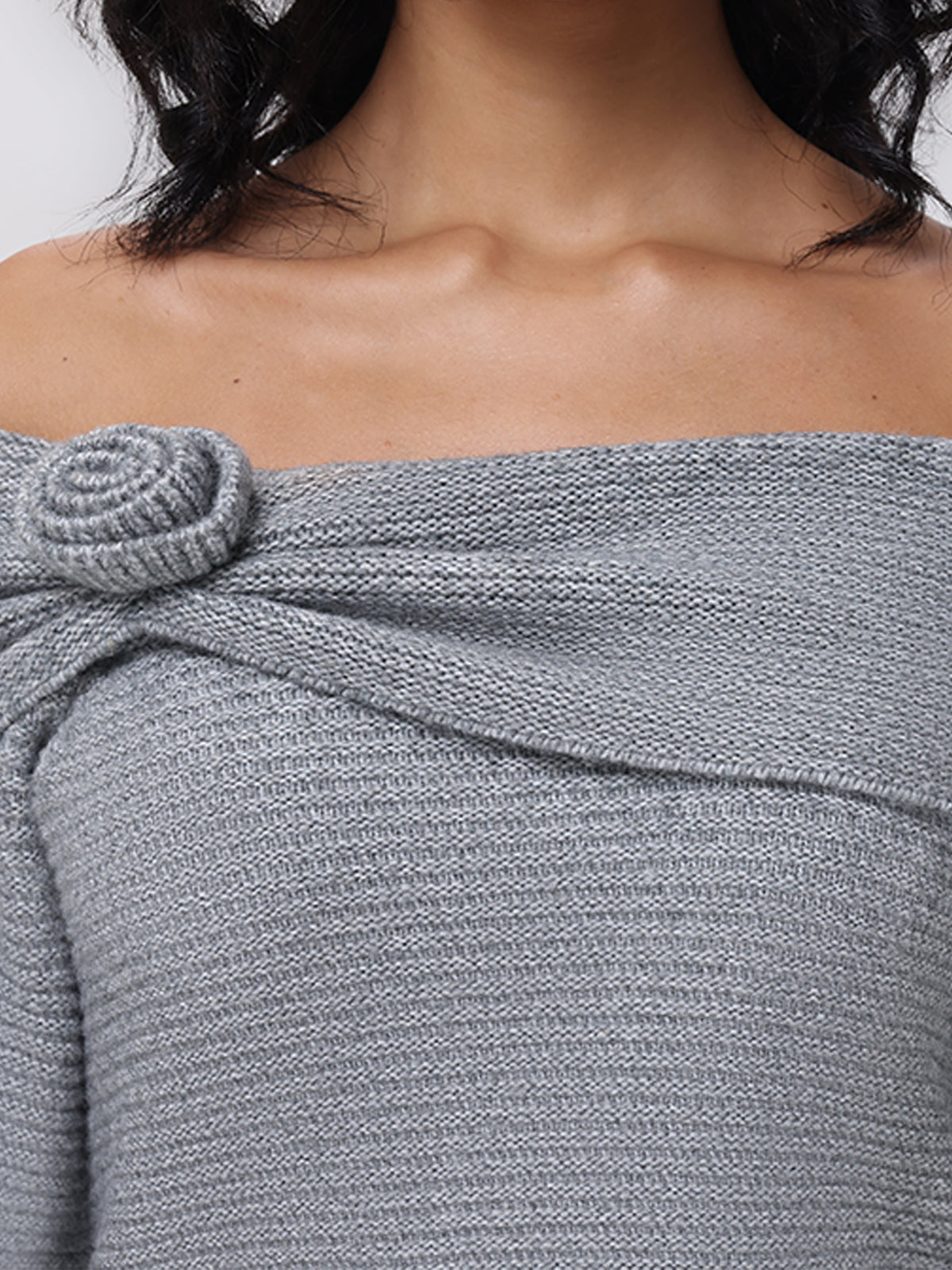 A model presents the 525 Americas Elizabeth Sweater, featuring an off-the-shoulder design in light gray knit with a delicate rosette accent on the left side. The image, focusing on the upper torso, highlights its intricate texture.