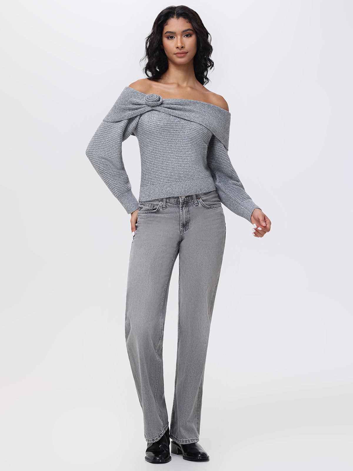 A person models a 525 America Elizabeth Sweater featuring an off-the-shoulder cut and decorative rosette, teamed with gray jeans. They stand neutrally against a plain white backdrop, arms at their sides, wearing black shoes.