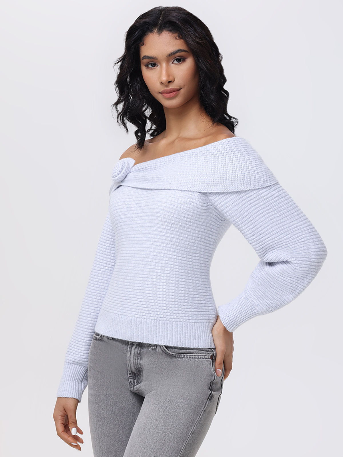 Against a plain backdrop, a person with medium dark hair wears a 525 America Elizabeth Sweater featuring an off-the-shoulder design, paired with gray jeans. One hand rests on their hip while the other hangs by their side.