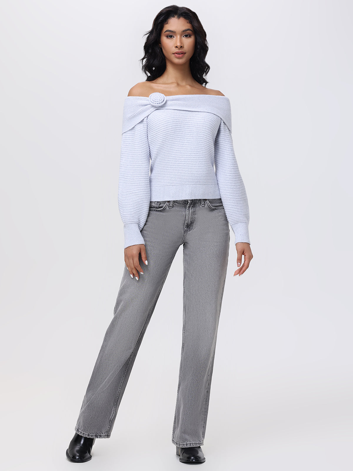 A person is standing in a relaxed pose, dressed in the Elizabeth Sweater from 525 America, featuring a light gray off-the-shoulder design with a rosette accent on the side. Its paired with gray jeans and black shoes, set against a plain light gray background.