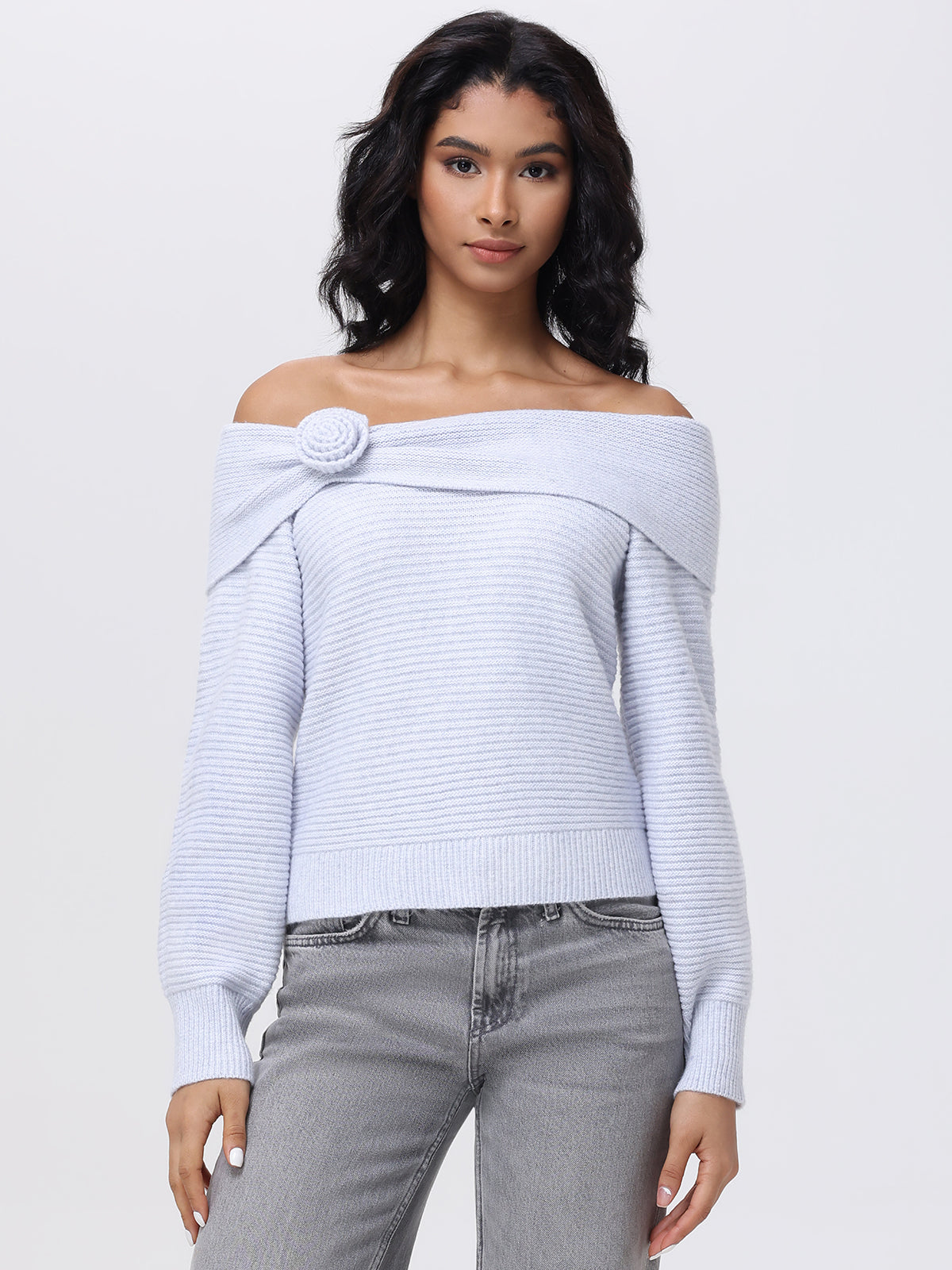 A woman with long dark hair is elegantly dressed in a light-colored 525 America sweater featuring a button detail on the collar and an off-the-shoulder design. She completes her look with light gray jeans against a plain white backdrop.