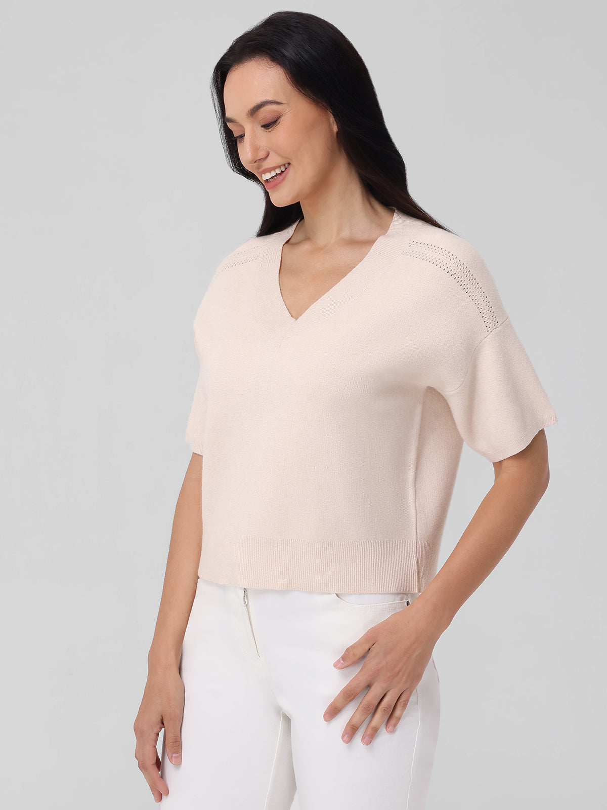 Wearing a light-colored V-neck short-sleeve knitted jumper by 525 America and white pants, an individual with long dark hair stands against a neutral backdrop, smiling subtly with timeless elegance as their right hand rests on their hip.