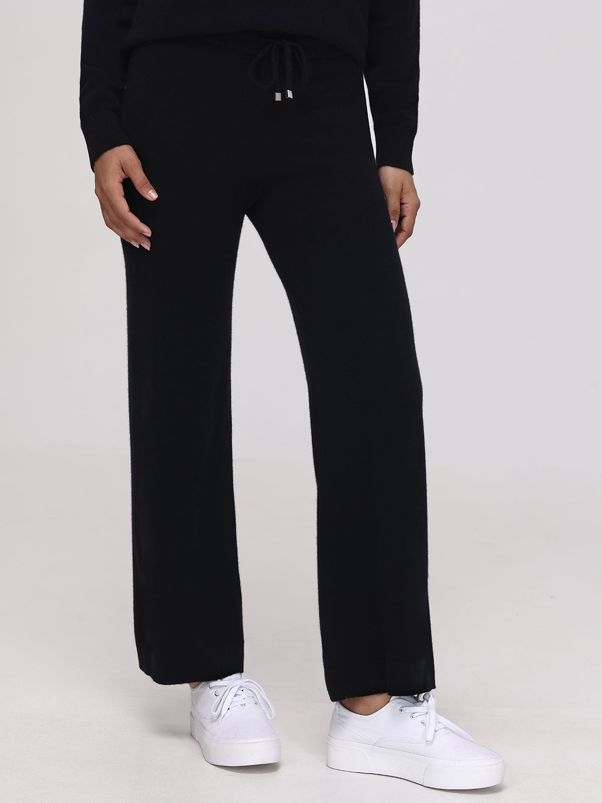 Against a plain background, only the lower half of a person is visible, showcasing Willow: Cashmere Straight Leg Pants in black. These luxurious drawstring trousers feature a relaxed fit paired elegantly with white sneakers.
