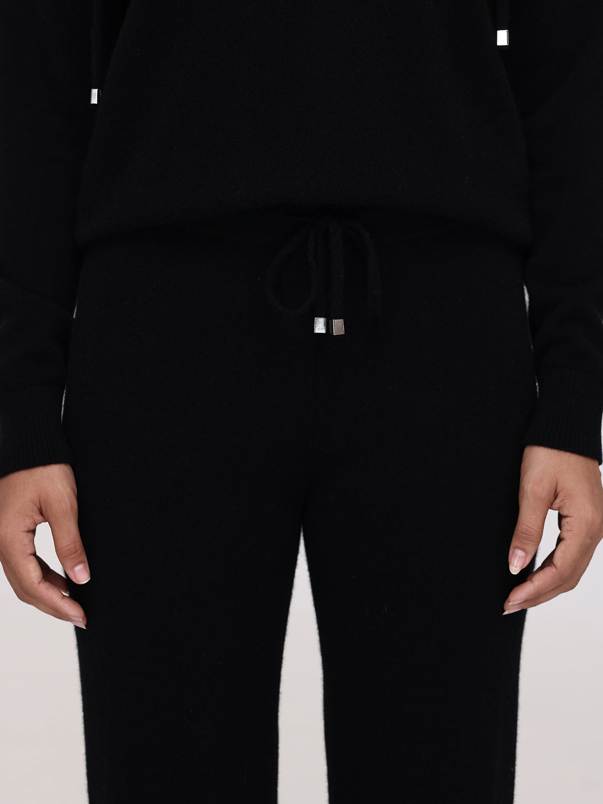A person wearing black cashmere straight-leg pants with a drawstring stands against a plain background, highlighting the texture and fit of the Willow Cashmere Pants. The relaxed hands add to the luxurious fashion appeal.