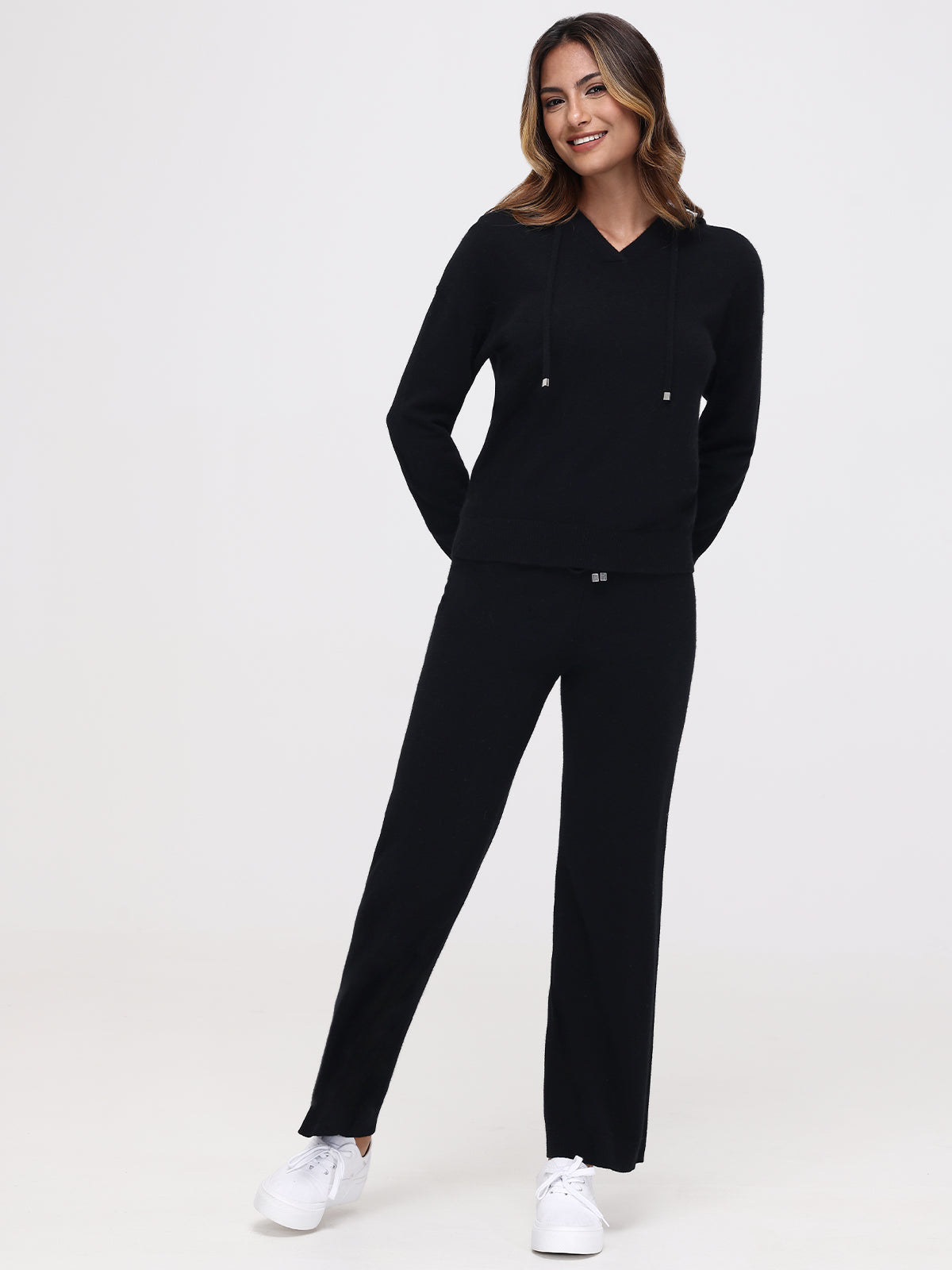 A person stands smiling in a black hooded sweater and Willows Cashmere Straight Leg Pants, with long hair and white sneakers against a plain white background, exuding an effortlessly chic vibe.