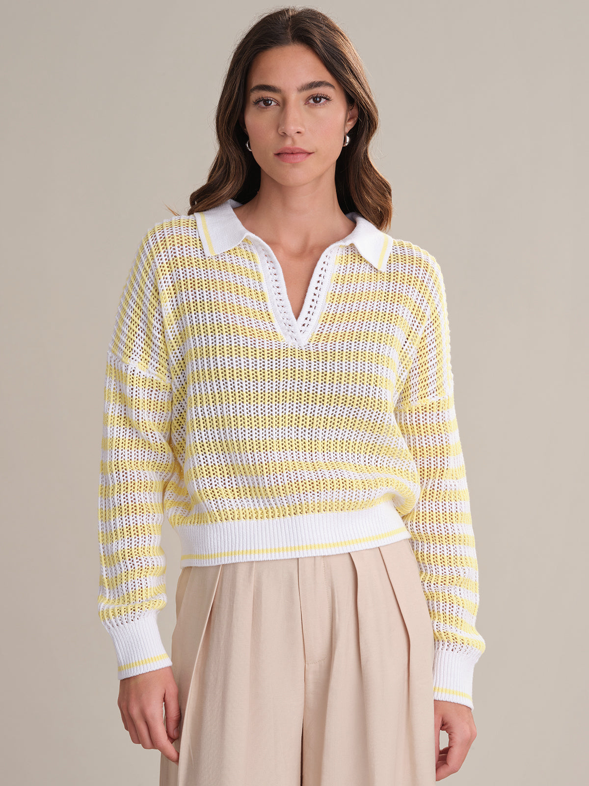 A woman with long brown hair models the 525 America Kimberly Stripe Polo Sweater, made from 100% cotton with a white collar, paired with beige high-waisted pants against a simple light background.
