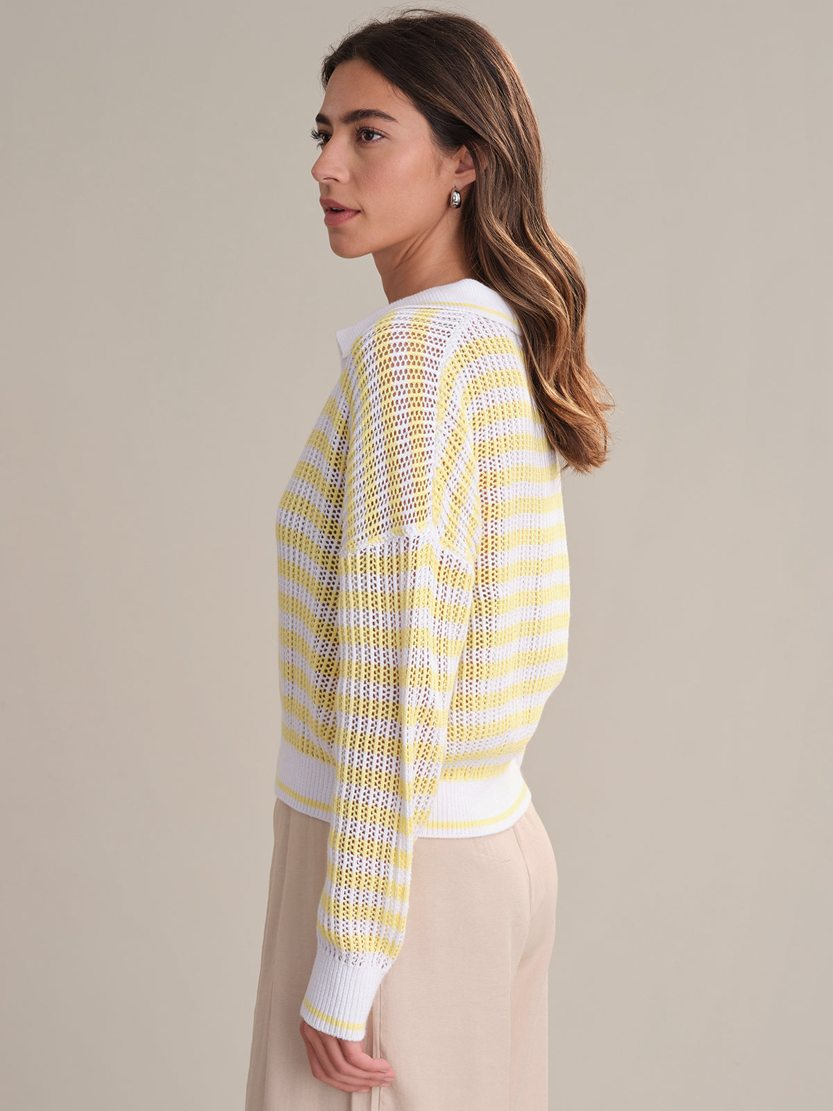 A woman with long brown hair stands sideways against a plain beige background, wearing 525 Americas Kimberly Stripe Polo Sweater in 100% cotton, paired with beige pants.