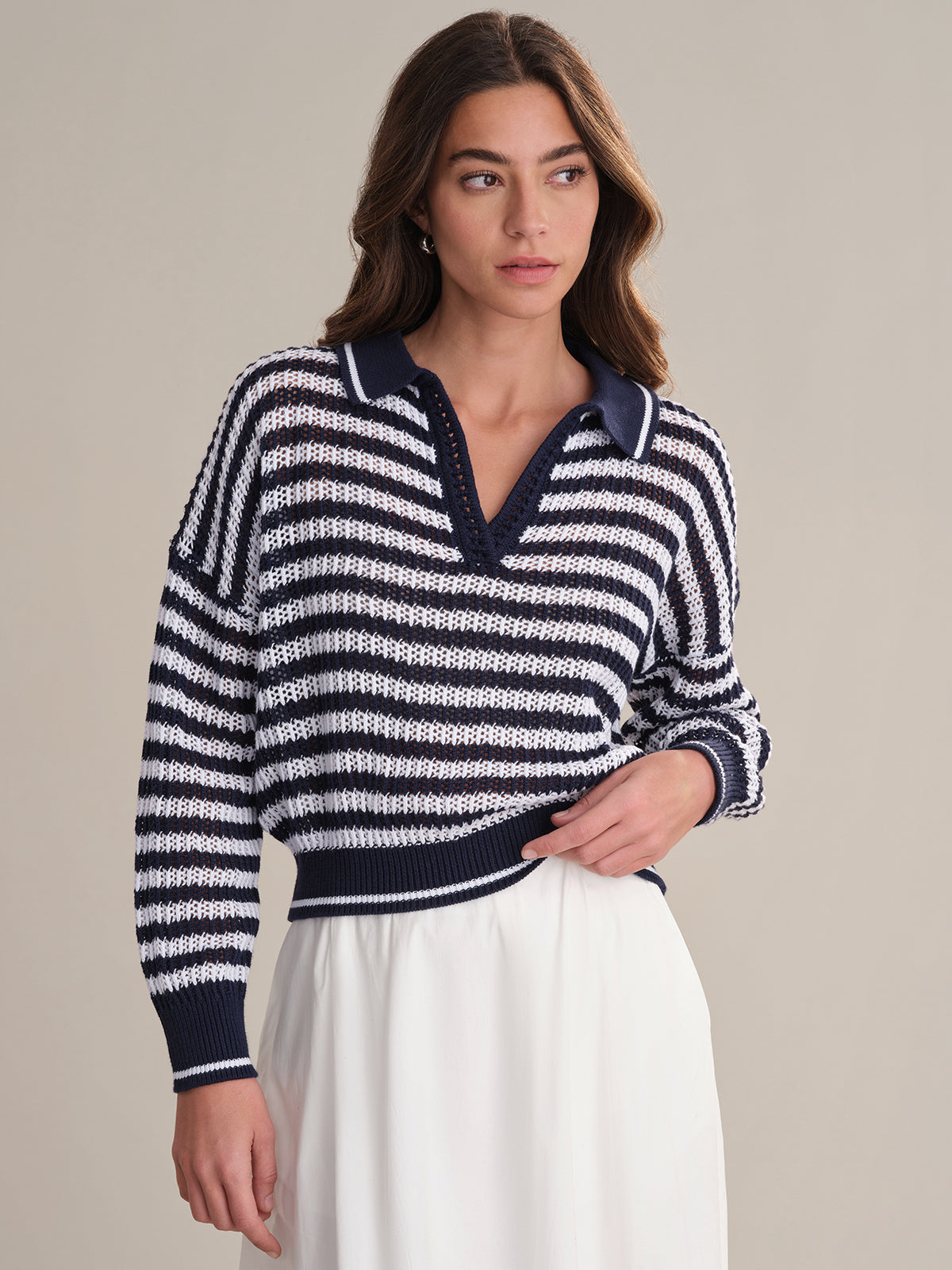 A woman with long brown hair models the 525 America Kimberly Stripe Polo Sweater, a navy and white cotton polo, paired with a white skirt against a plain beige background as she looks slightly to the side.